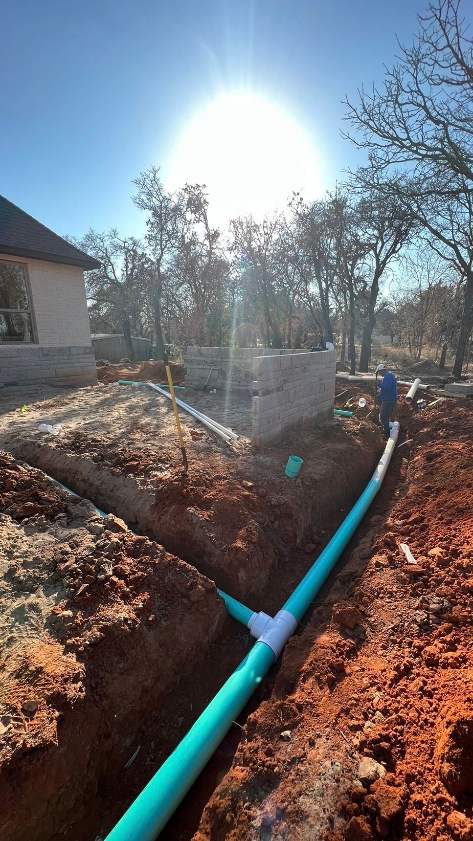 A turquoise pipe runs through a dirt trench beside a house, with bright sunlight and bare trees.