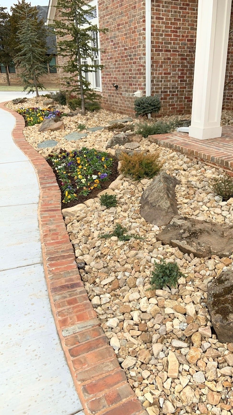 Brick-edged rock garden with small shrubs and flowers beside a house walkway