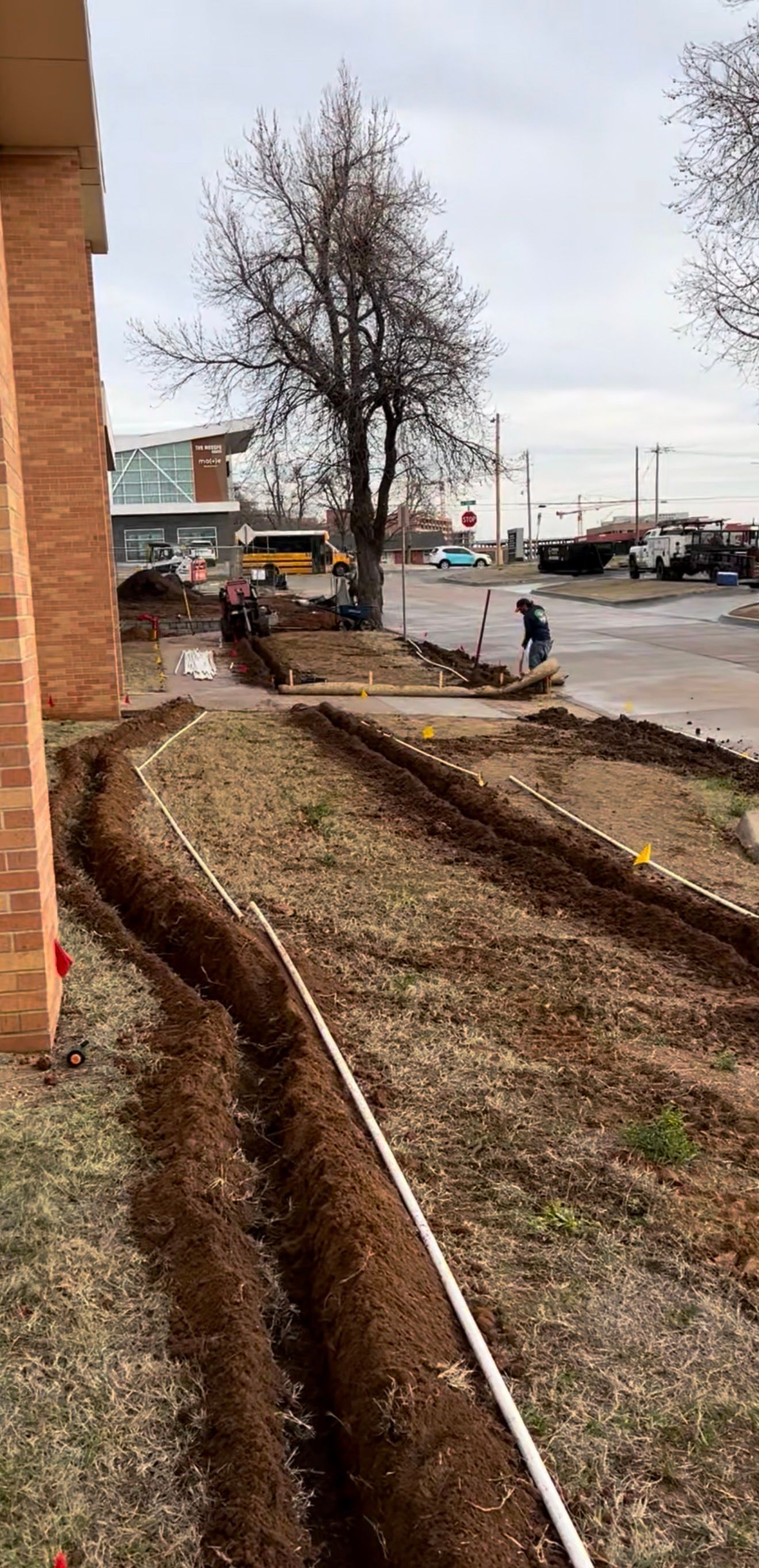 Narrow landscaped strip beside a brick building and wet street, with bare trees and a few workers in the distance.