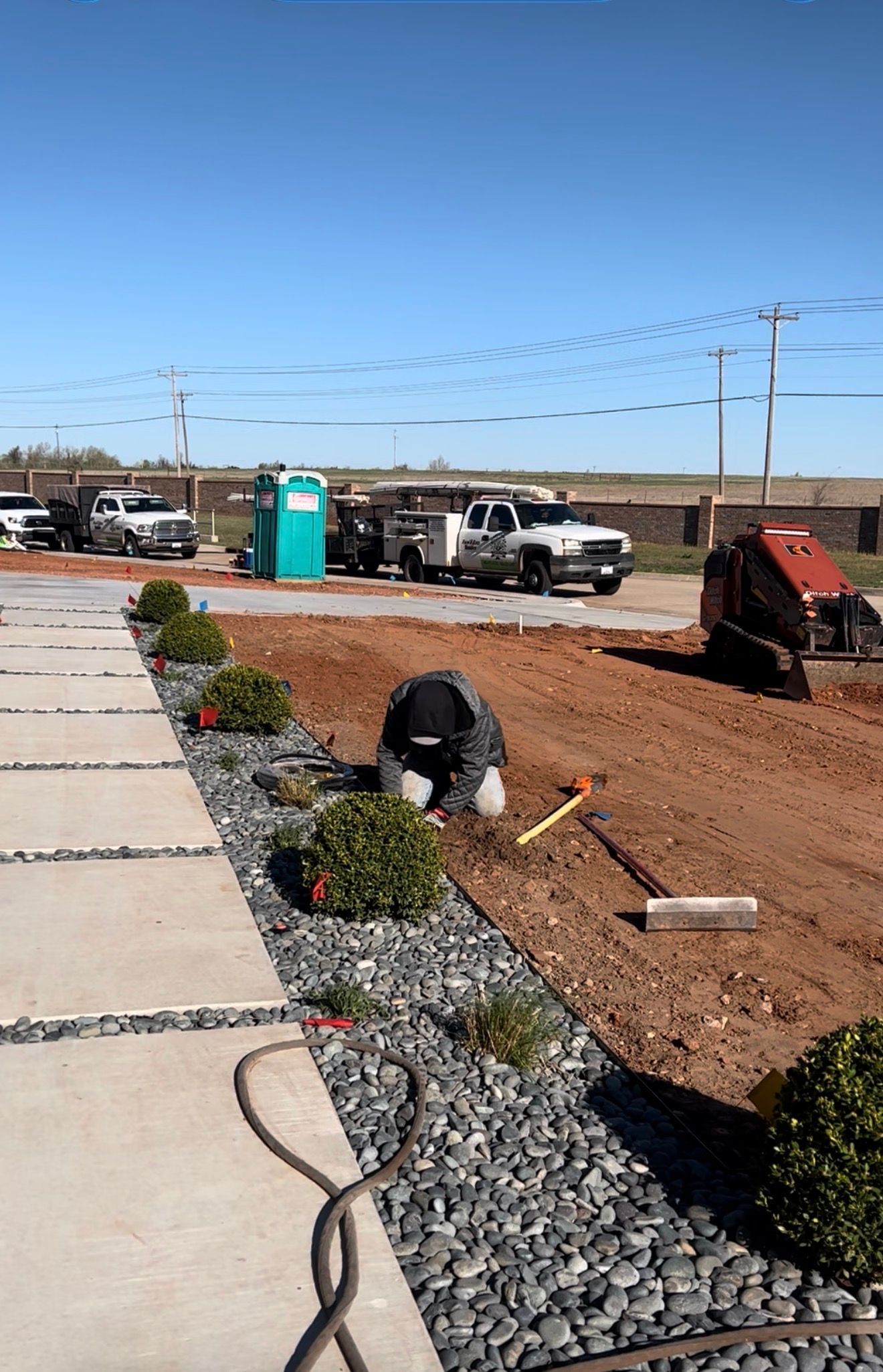Roadside construction site with sidewalk, gravel landscaping, workers, equipment, and a portable toilet under a clear sky