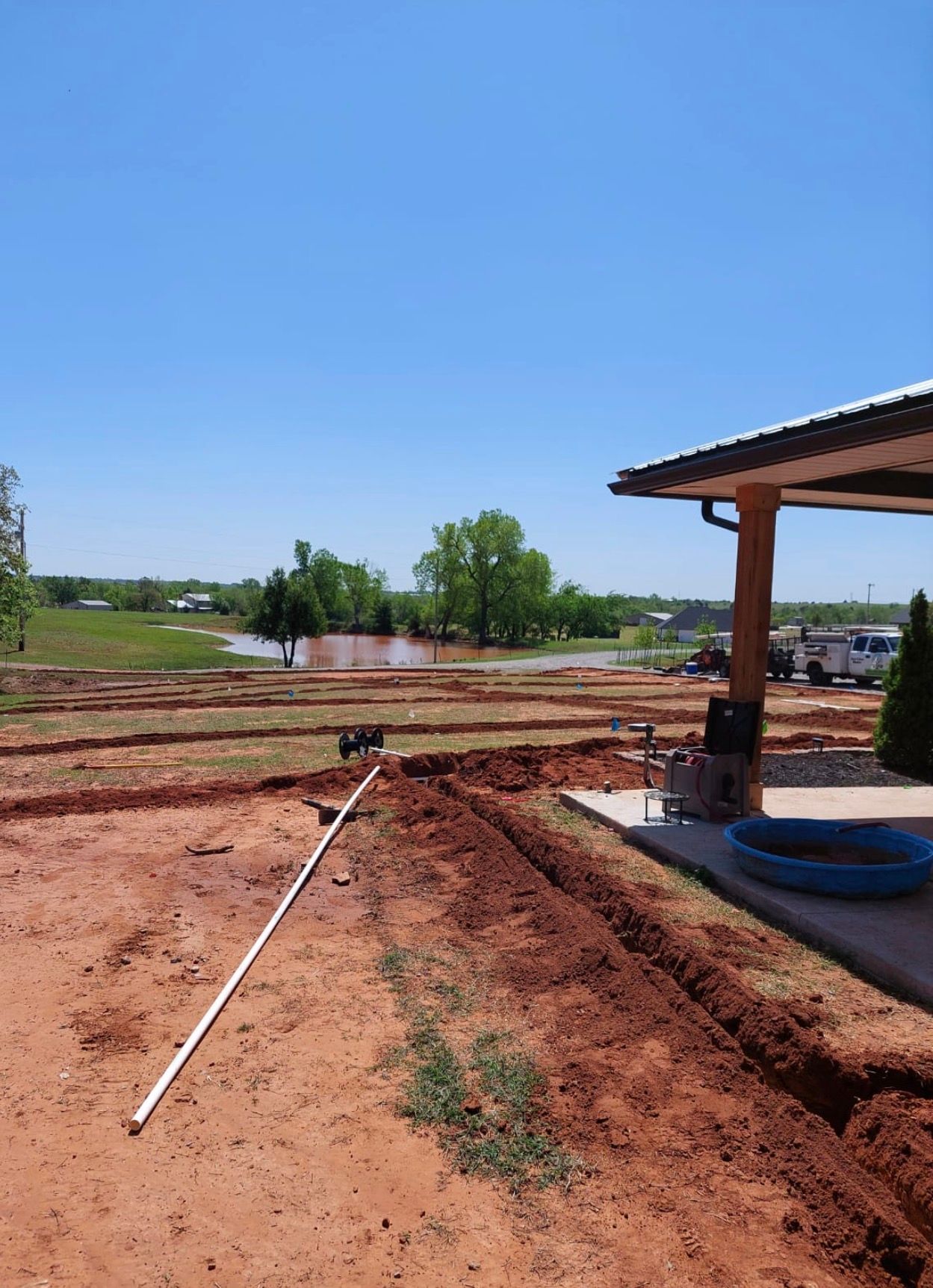 Rural construction site with a dirt trench, white pipe, and a partially roofed structure under a clear blue sky