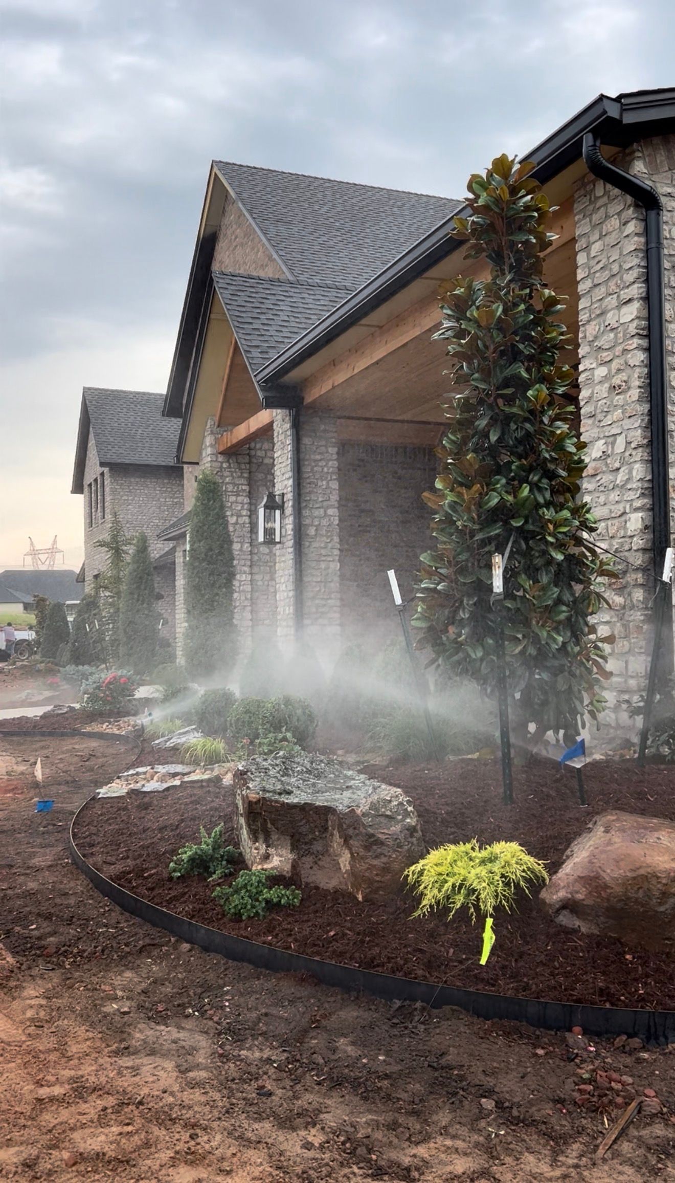 Sprinklers mist a landscaped garden beside a stone house at dusk