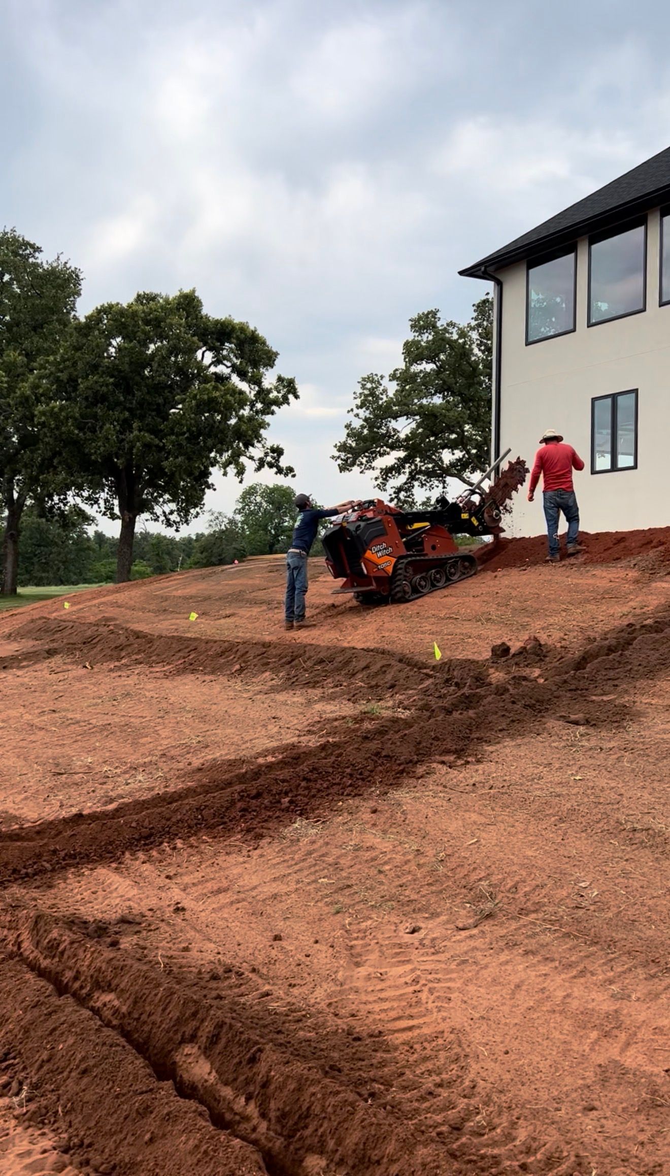 Workers operating a red tiller on a dirt slope beside a house under cloudy sky