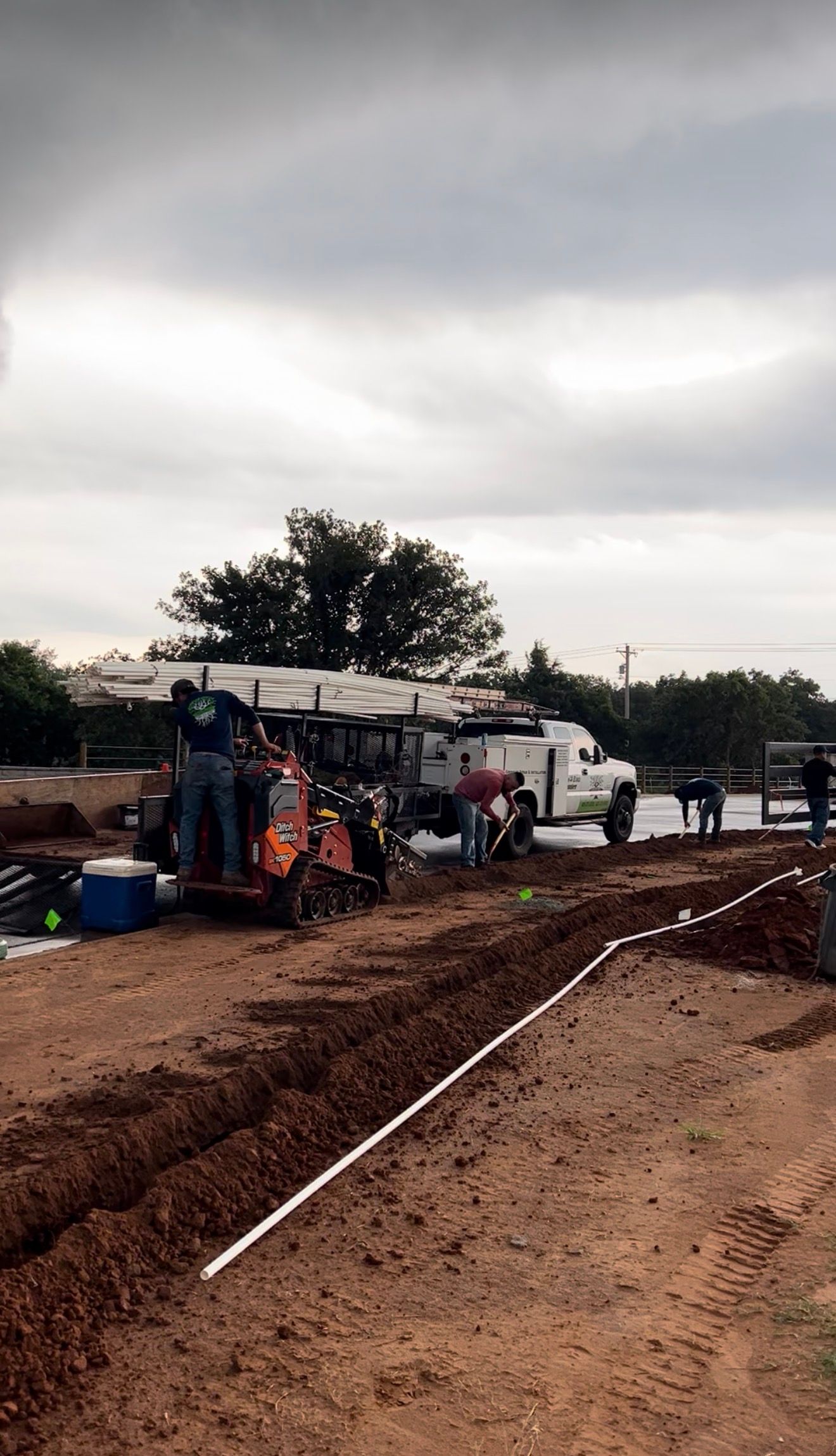 Workers paving a muddy road beside parked trucks under a cloudy sky