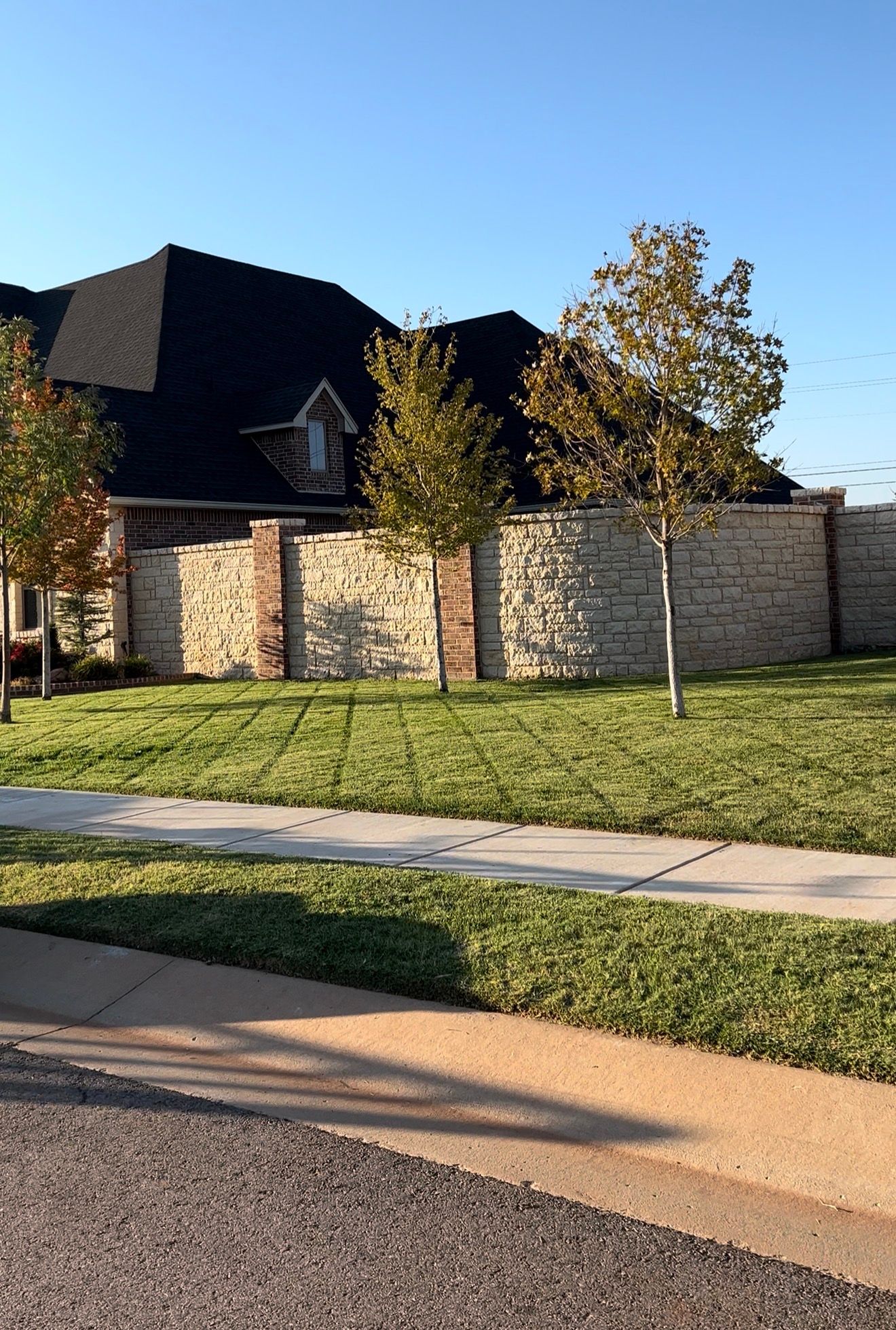 Residential street with stone wall, young trees, and a large house behind a grassy lawn under a clear blue sky