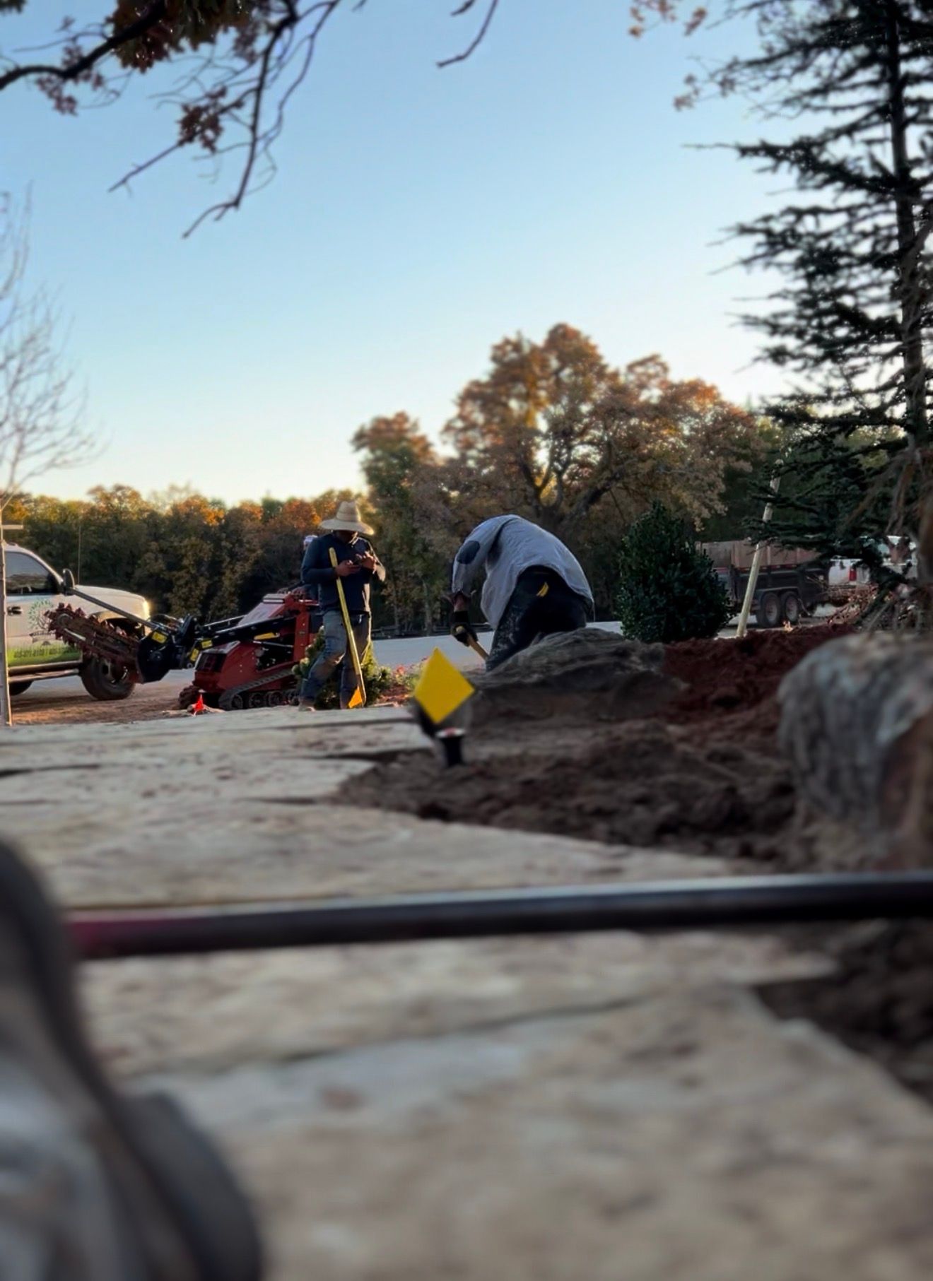 Workers with tools at a roadside excavation site at dusk, with trees and equipment in the background