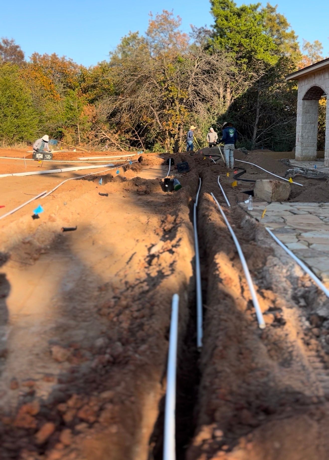 Trenched dirt construction site with PVC pipes laid beside a stone walkway and house, trees in background
