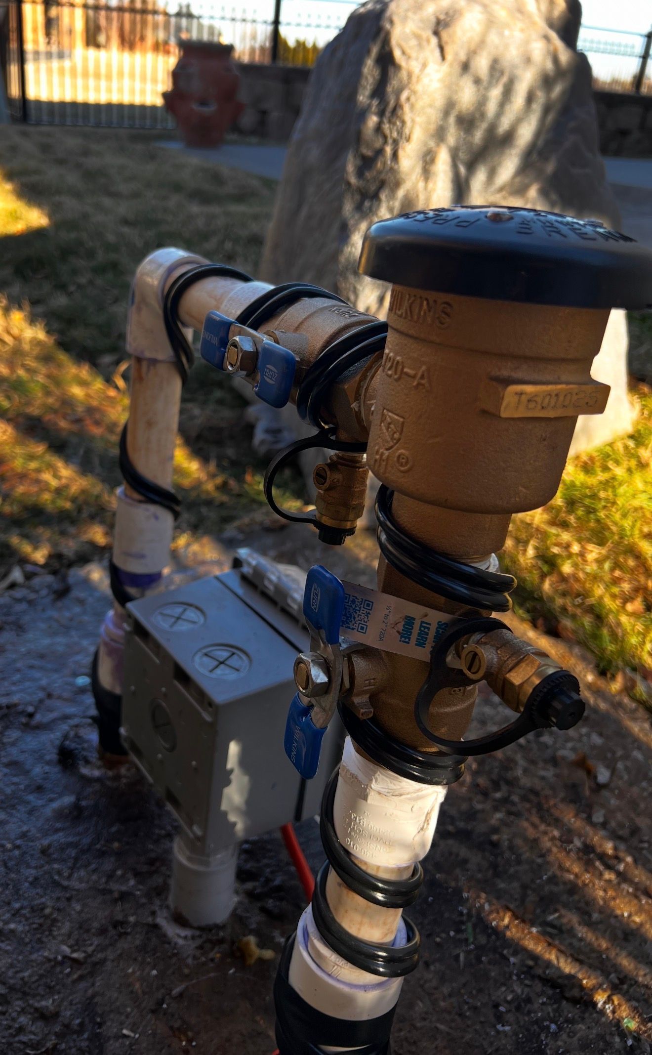 Close-up of an outdoor metal pipe assembly with a valve and insulation near grass and a tree trunk