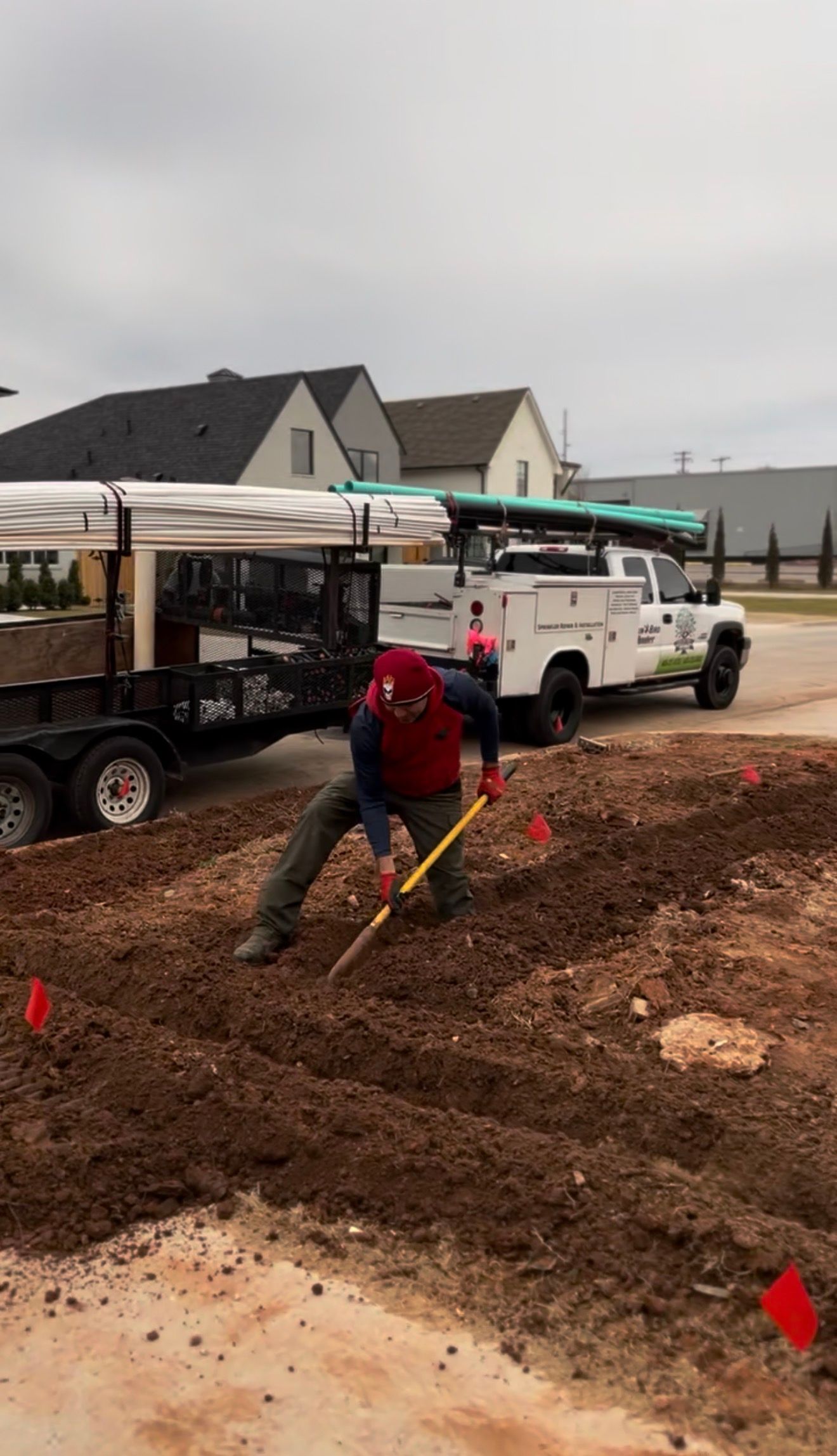 Worker in red jacket shoveling dirt beside utility trucks at a construction site.