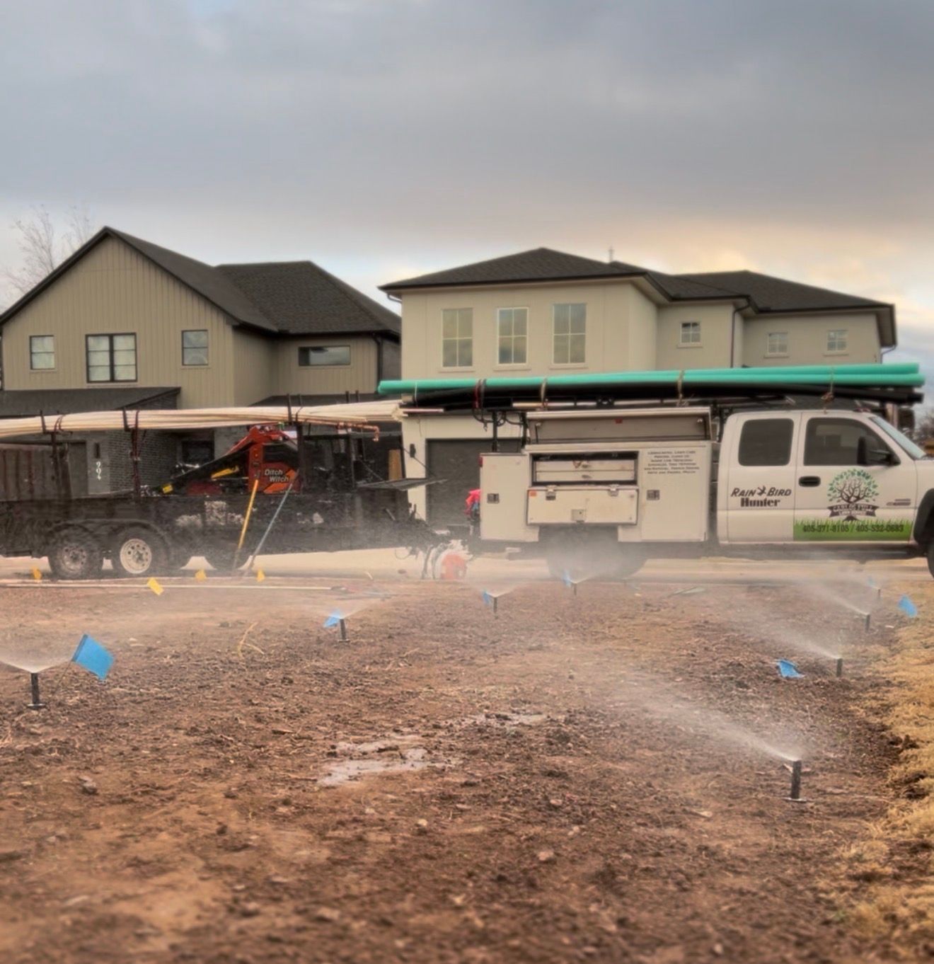 Irrigation system spraying water in a muddy lot in front of suburban houses with work trucks nearby