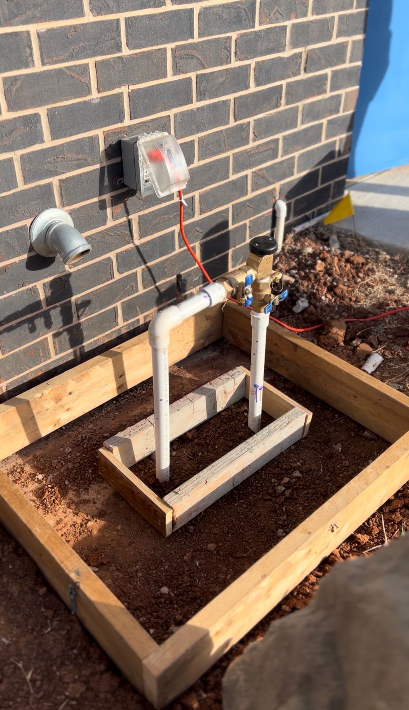 Outdoor plumbing pipes and valves in a wooden bed beside a brick wall, with a hose reel attached.
