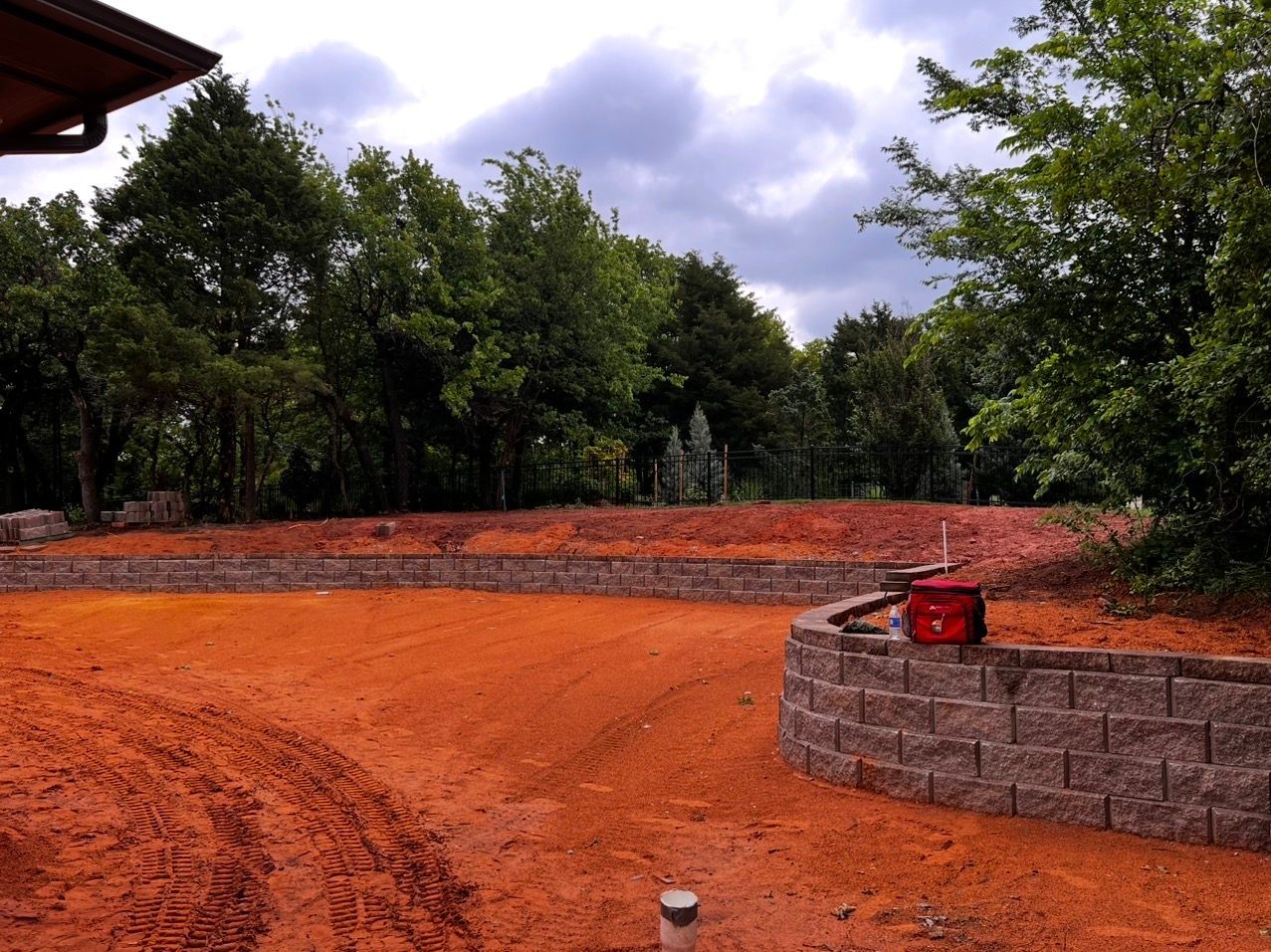 Reddish dirt courtyard beside a low brick wall, with trees and a cloudy sky in the background.