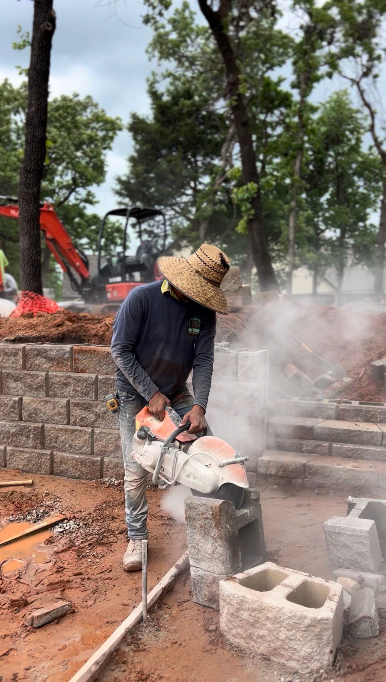 Worker cutting concrete blocks outdoors, with dust rising near trees and construction materials.