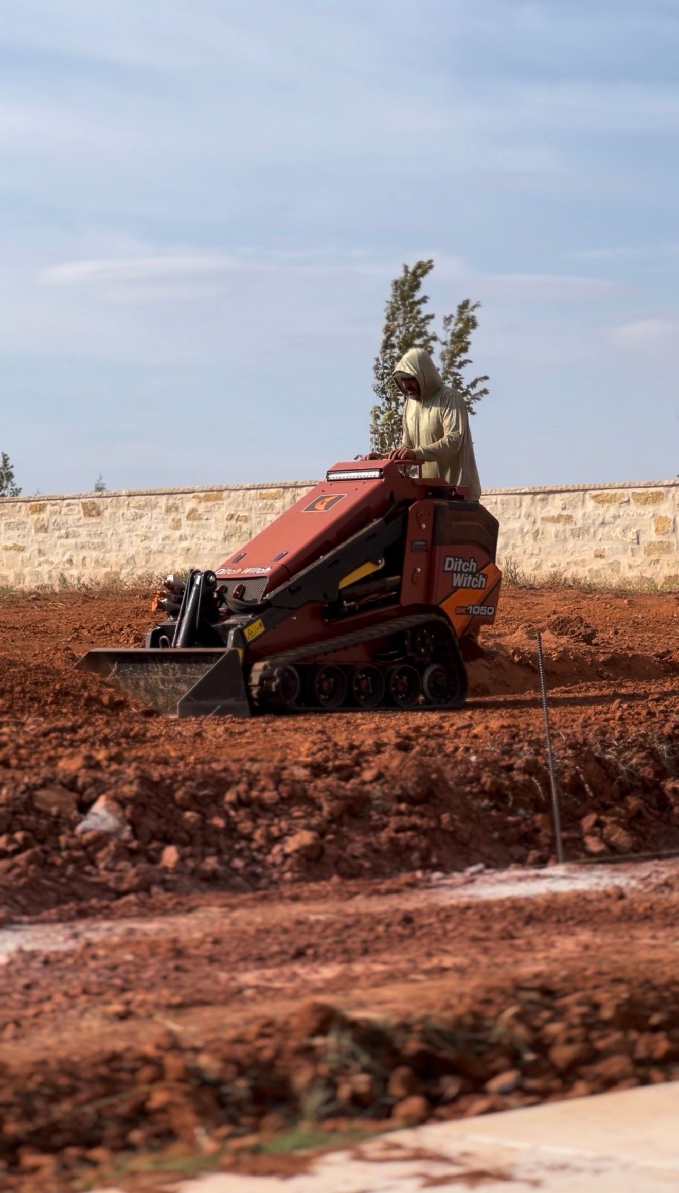 Red agricultural machine on a muddy field, spraying water into the air near a low wall under a blue sky