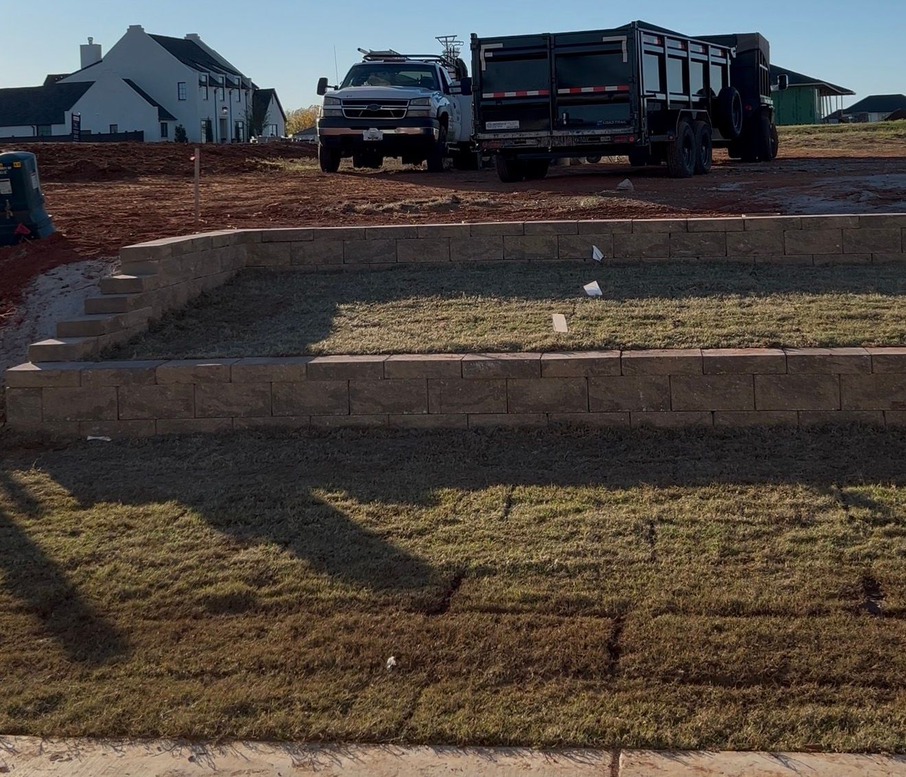 Construction site with two trucks, stacked retaining walls, and a patchy grassy foreground under a clear sky