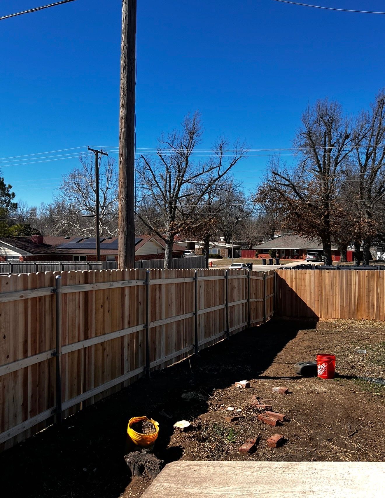 Backyard with wooden fence, dirt ground, and small red bucket under a clear blue sky.