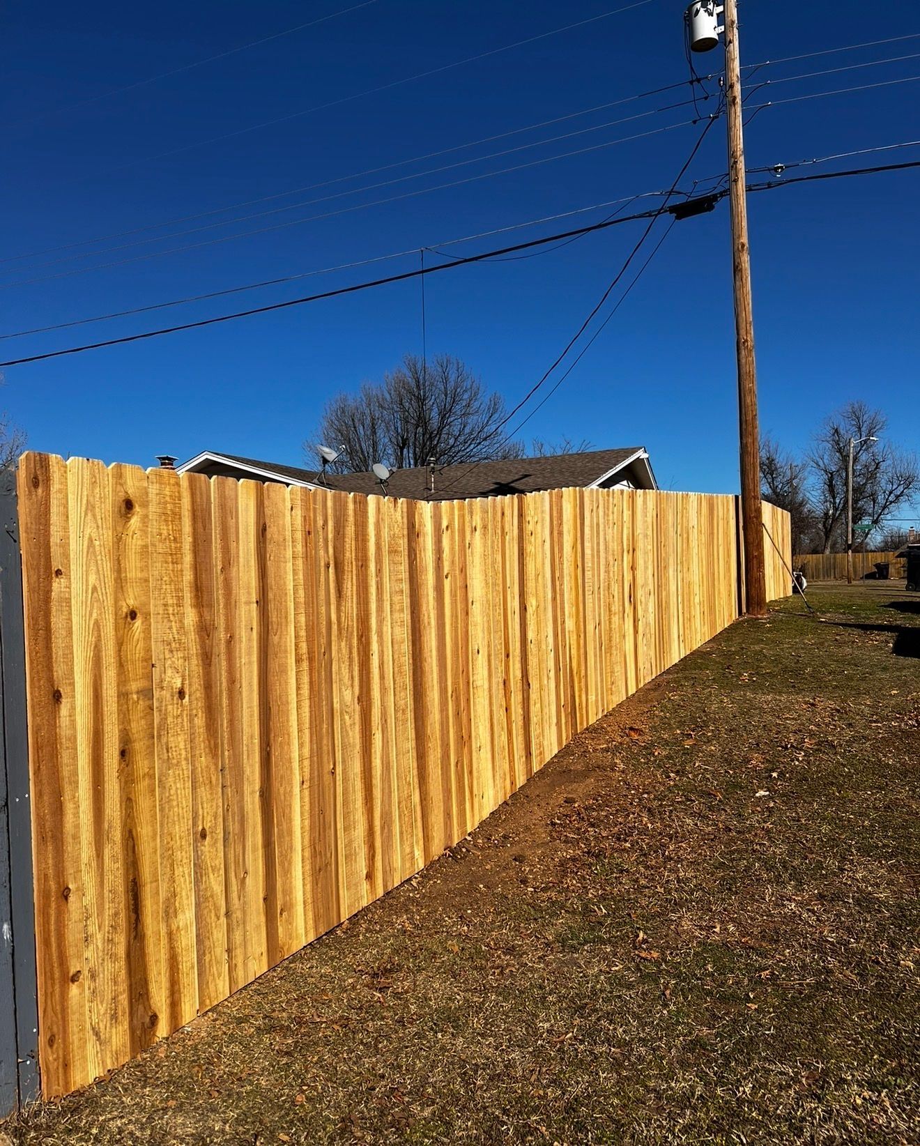 Wooden privacy fence along a grassy slope under a clear blue sky, with power lines overhead.