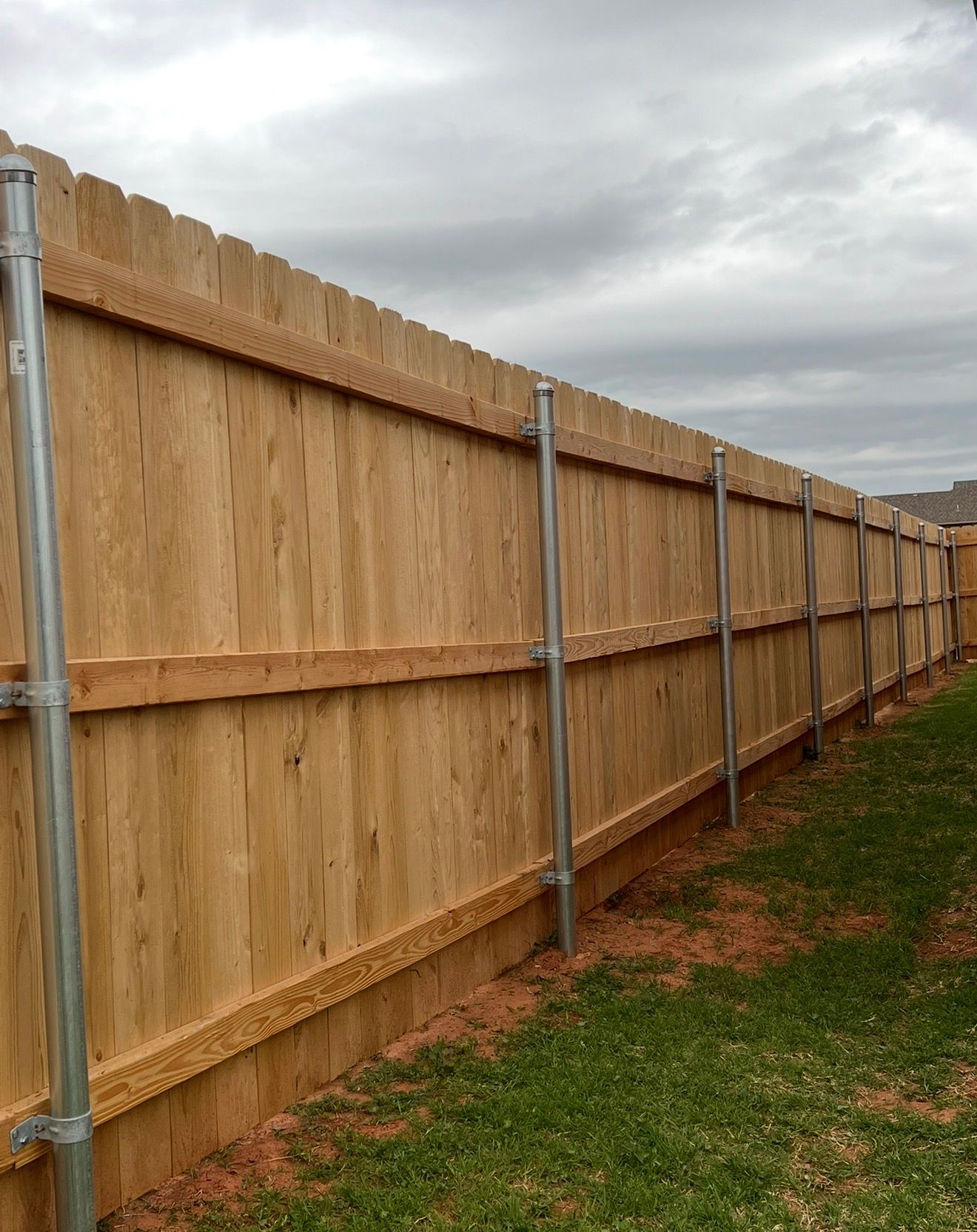 Wooden privacy fence with metal posts along a grassy yard under a cloudy sky