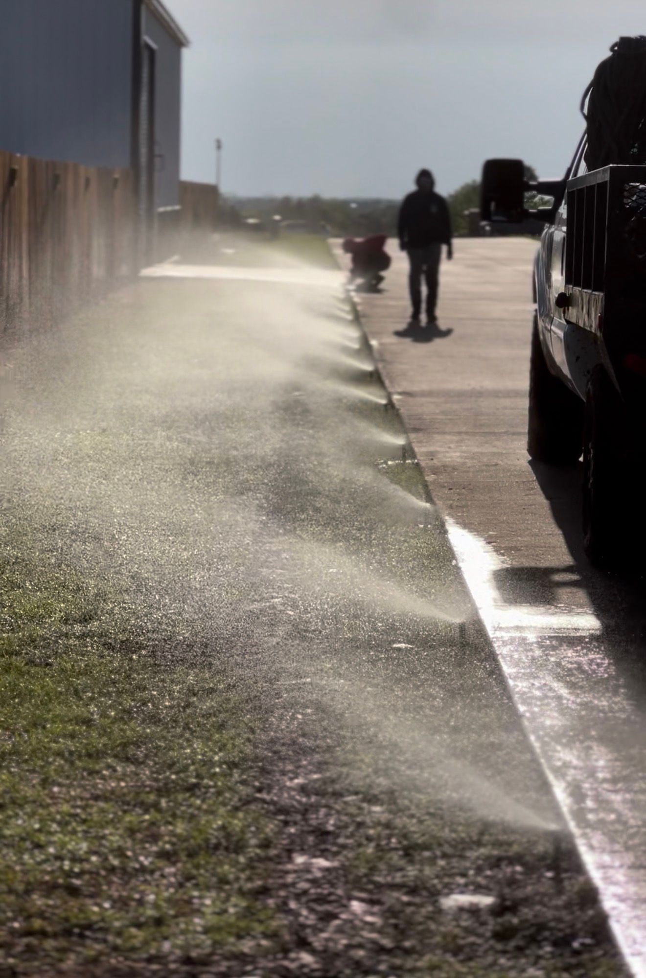 A child and adult walk beside a wet sidewalk with sprinkler mist along a building.