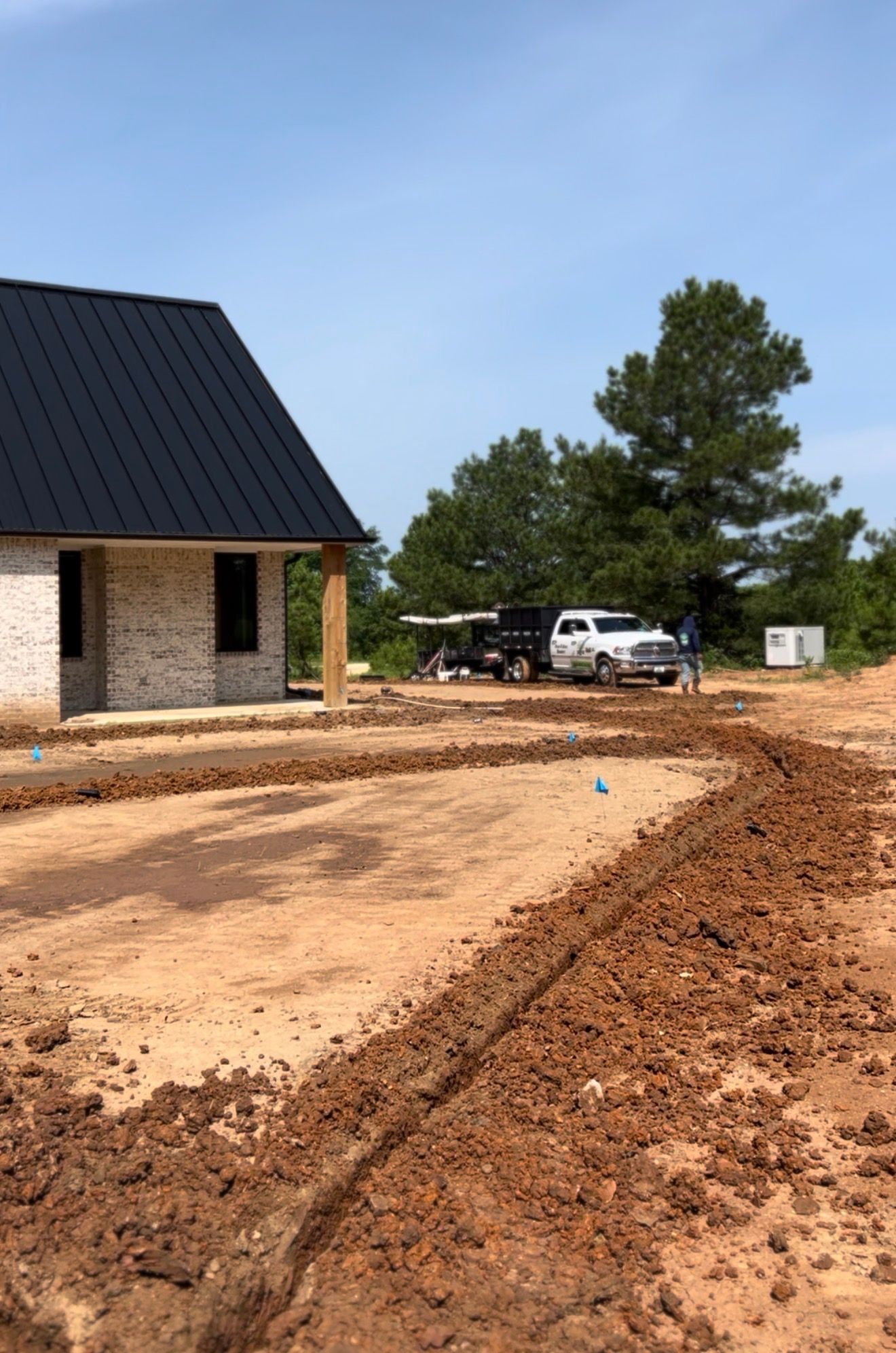 New house with dark roof beside a muddy dirt driveway and parked trucks under a blue sky