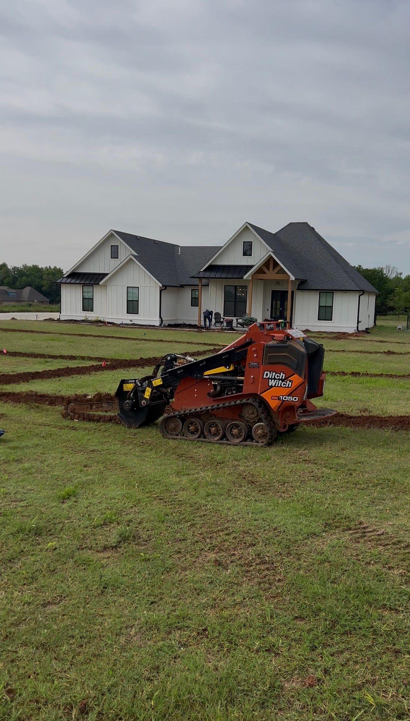 Orange skid steer on a muddy lawn in front of a white house under cloudy skies