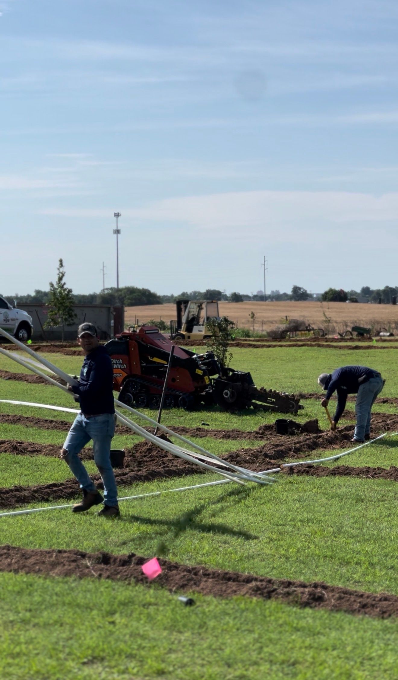 Workers digging a trench in a grassy field near farm equipment on a sunny day