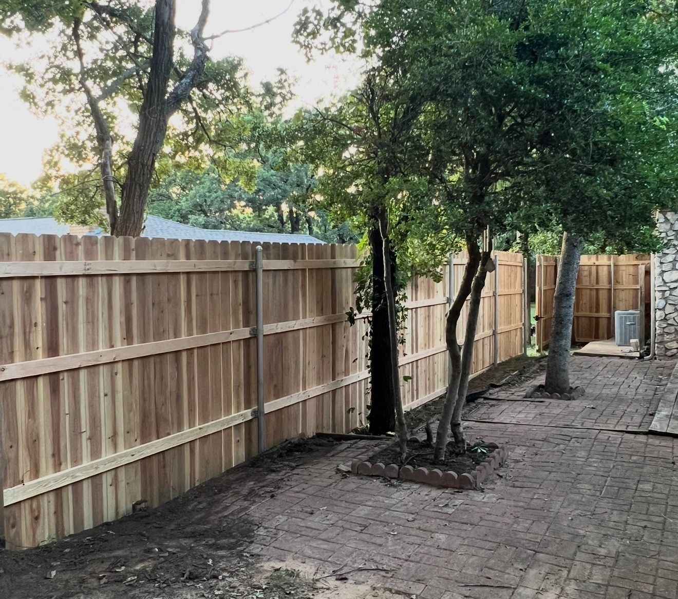 Wooden fence along a brick patio with trees and a small fenced tree planter in a backyard.