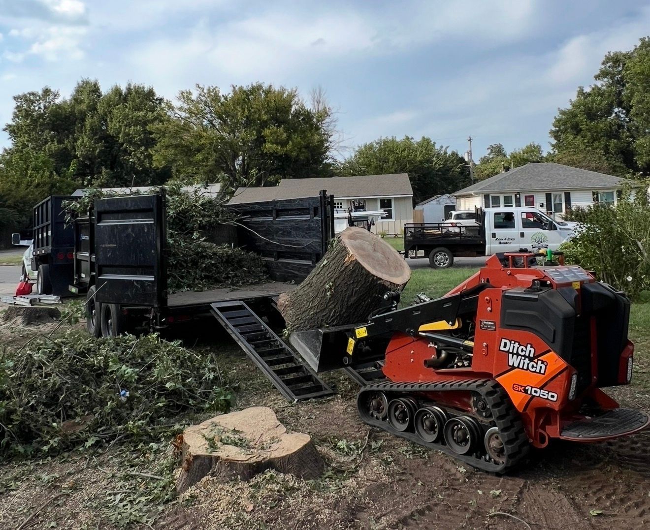 Orange skid steer loading a large tree trunk onto a truck in a suburban yard.