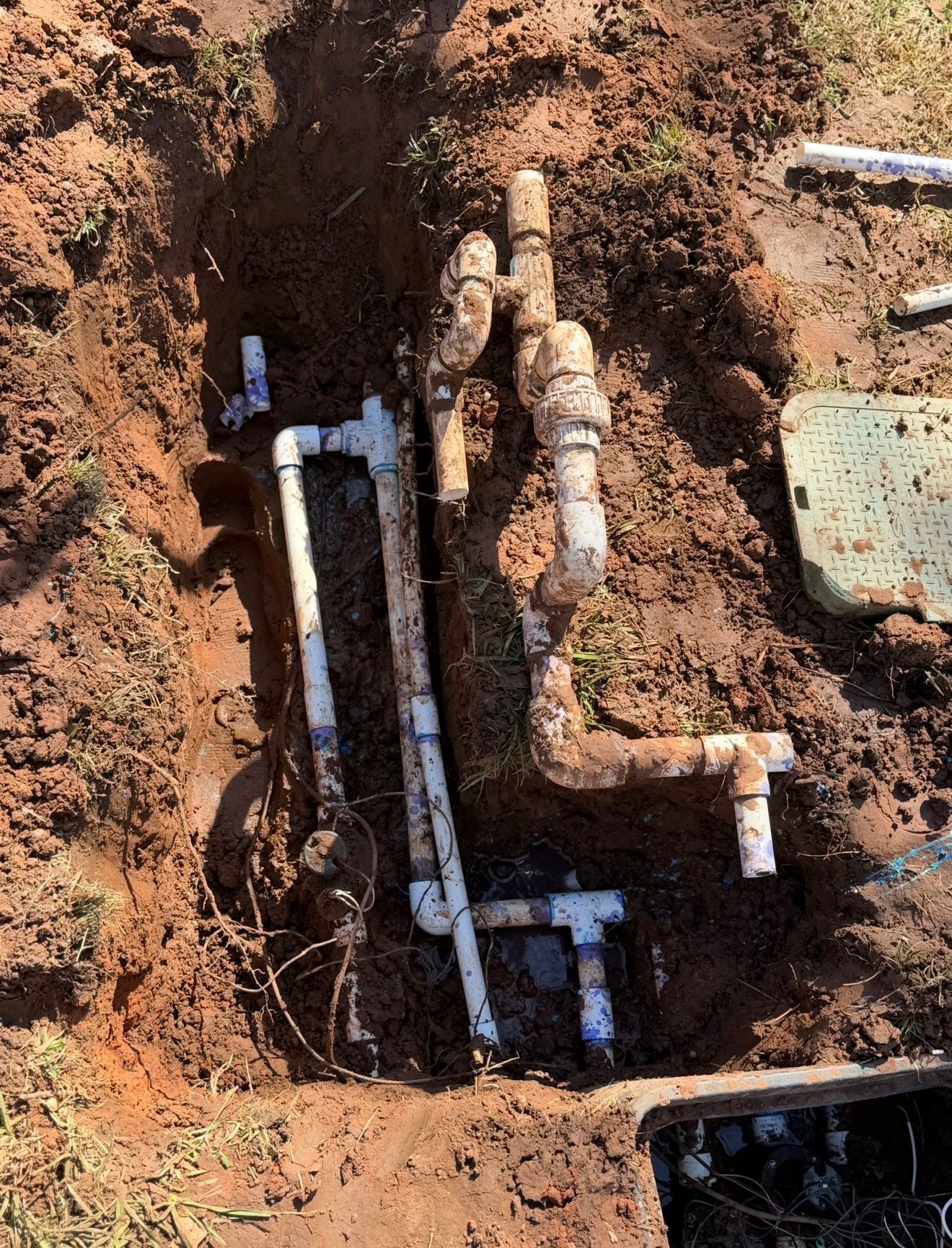 Open trench with exposed underground pipes and fittings in reddish dirt during utility repair
