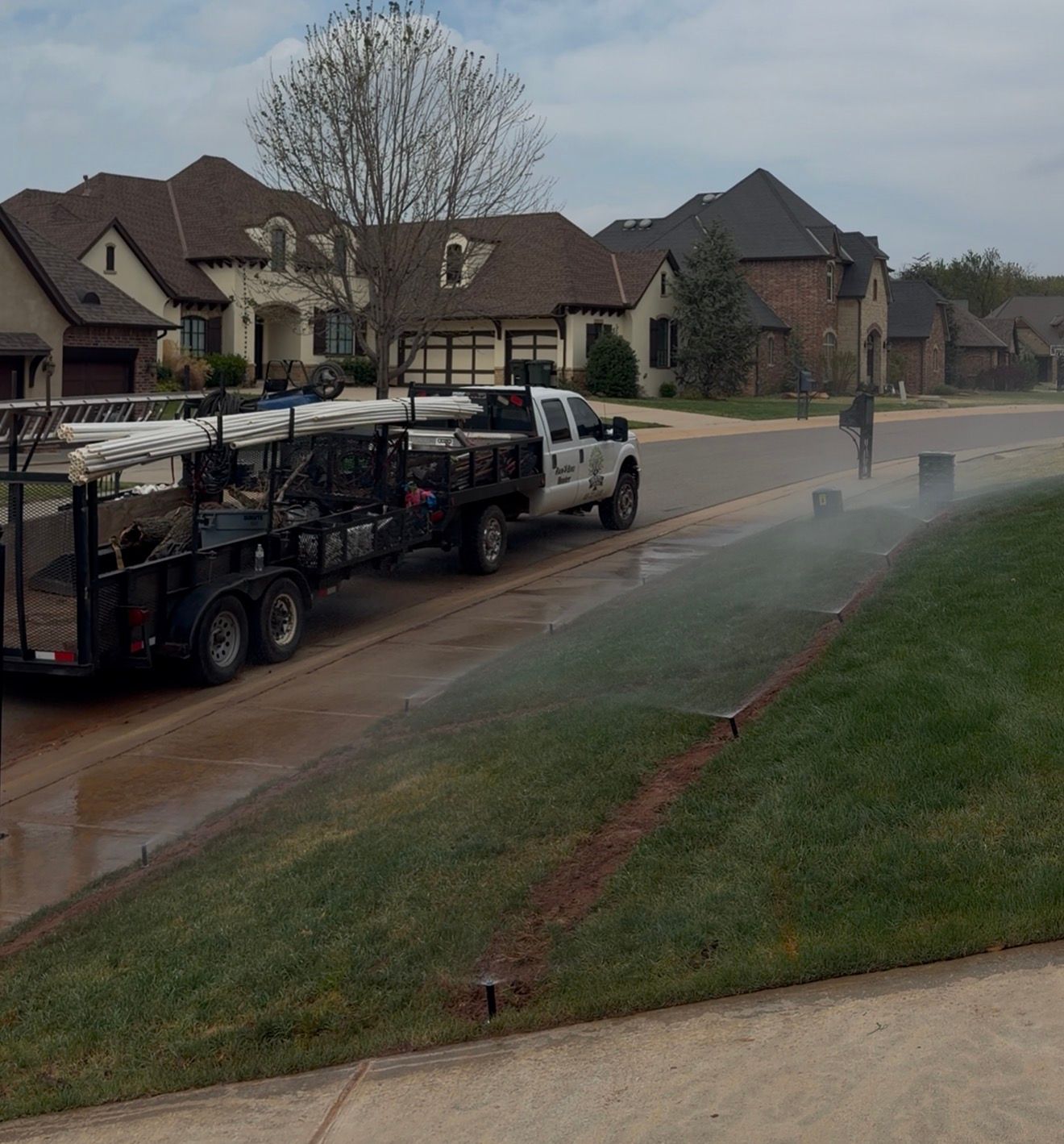 Landscape crew spraying water on a suburban street beside parked work truck and trailer.