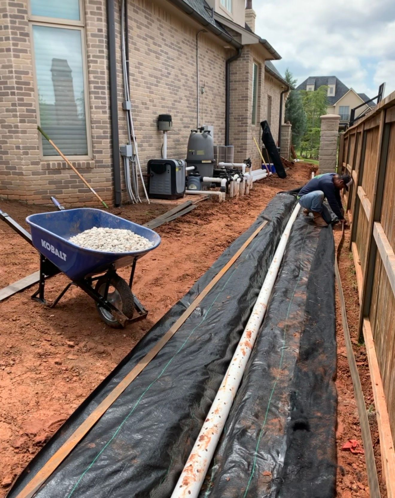 Worker laying black landscaping fabric beside a house, with wheelbarrow and red dirt path.