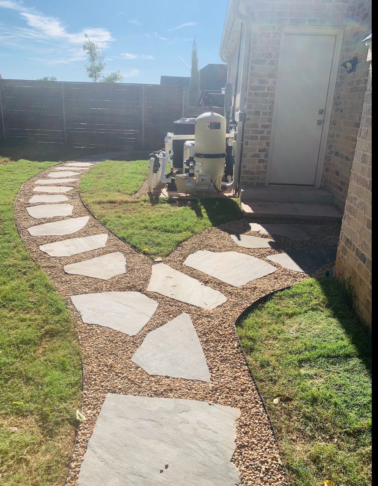 Curved stone walkway through a grassy yard beside a brick house and outdoor HVAC unit