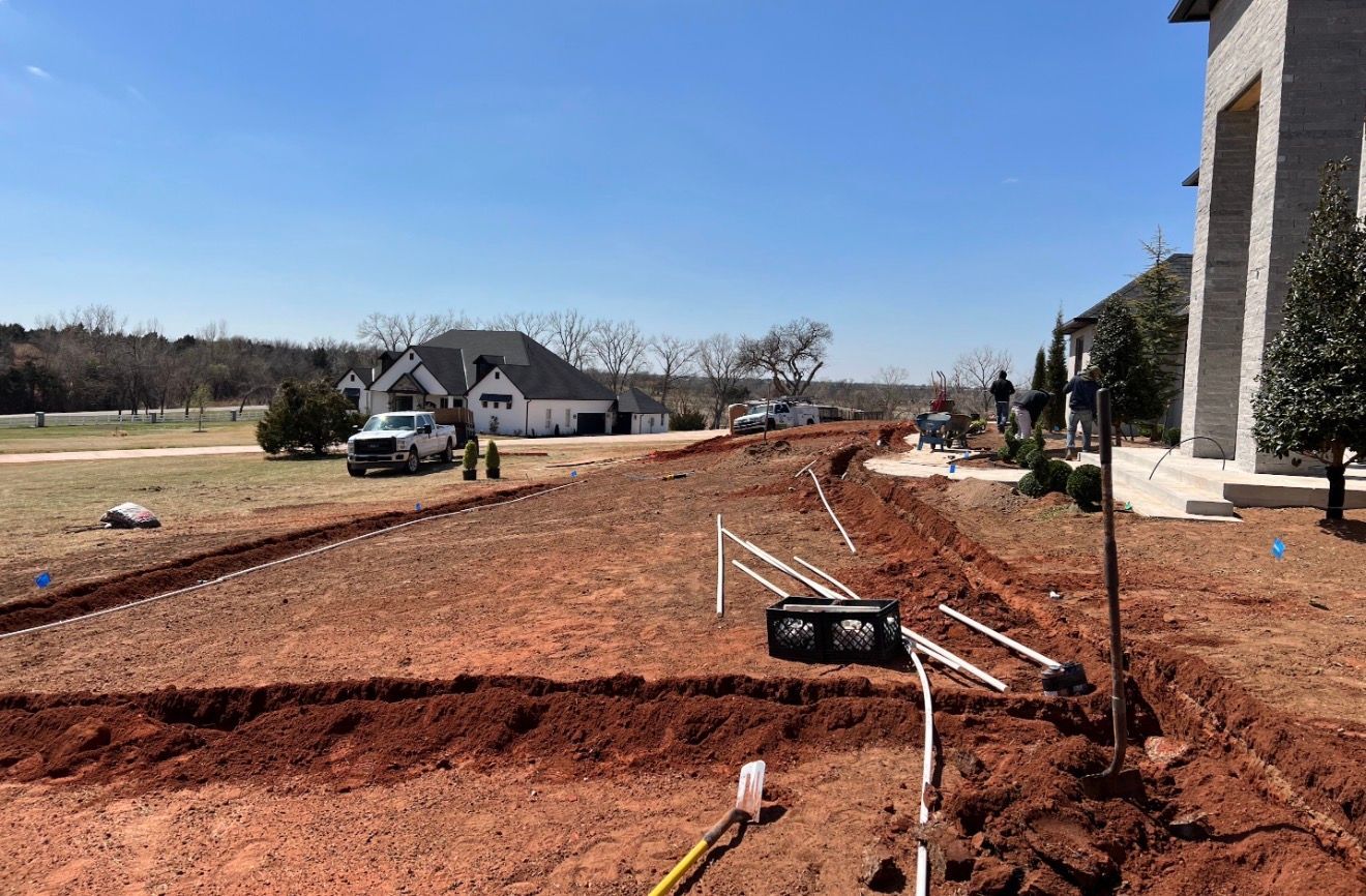 Construction site beside a house, with red dirt, pipes, and a truck on a rural road under a clear sky.