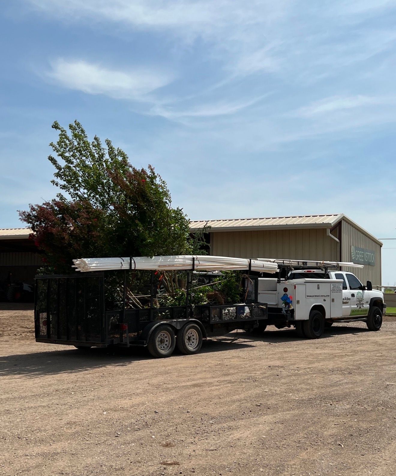 White truck hauling a trailer with potted trees beside a beige warehouse on a gravel lot