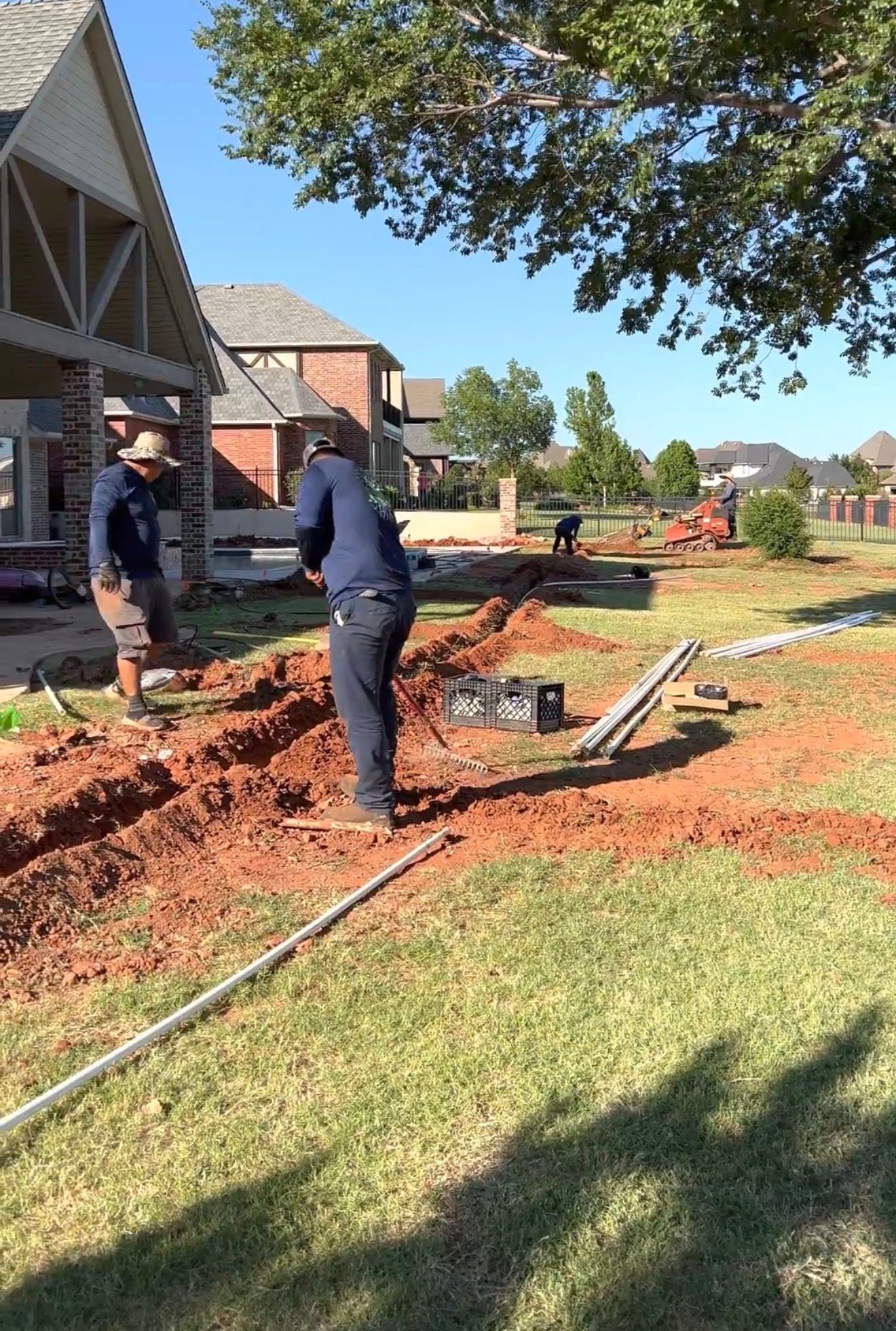Two workers repair a trench beside townhouses, with pipes and dirt scattered across the grassy yard.