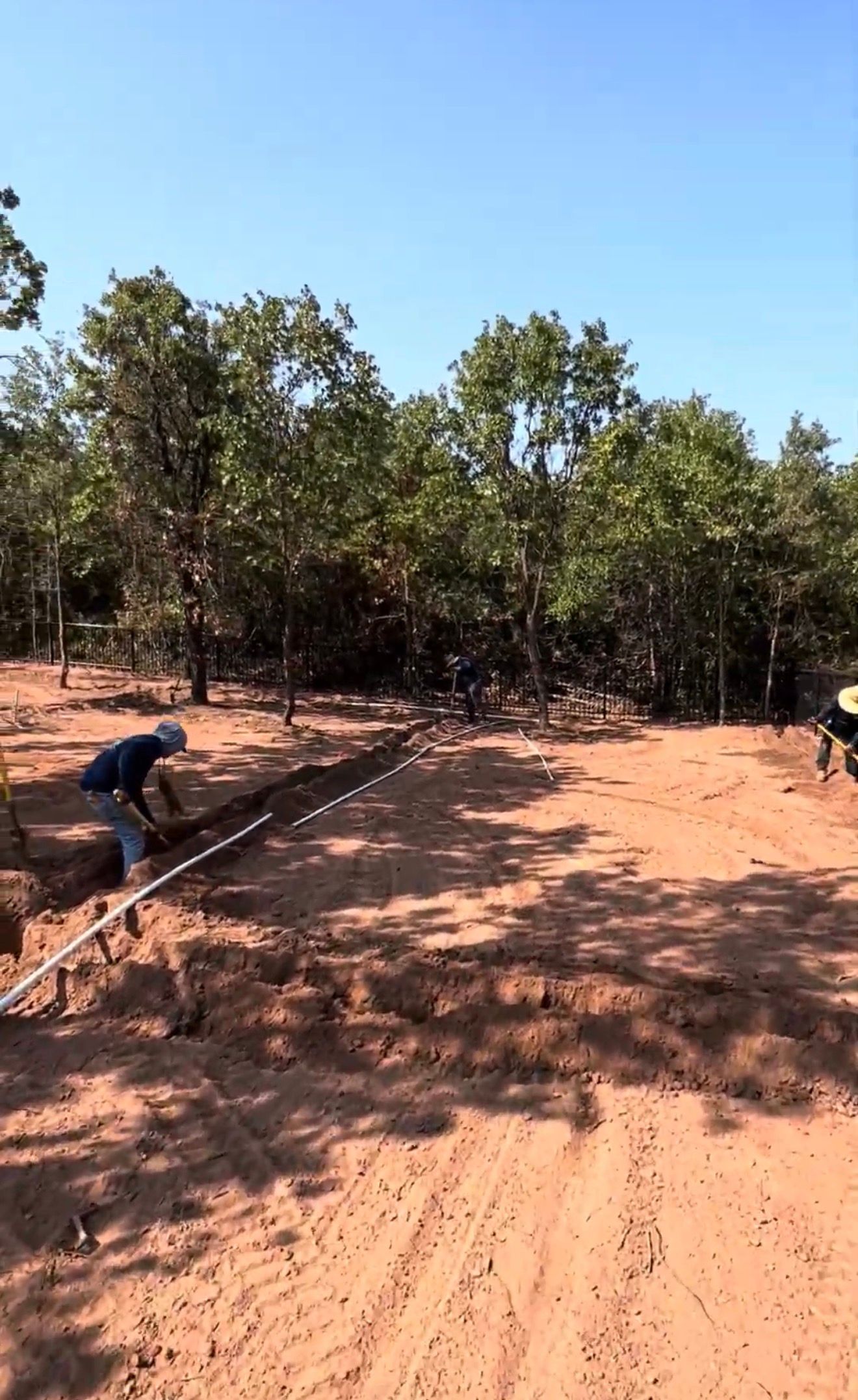 Person working on a dirt path beside a water hose in a dry, wooded area under a clear blue sky