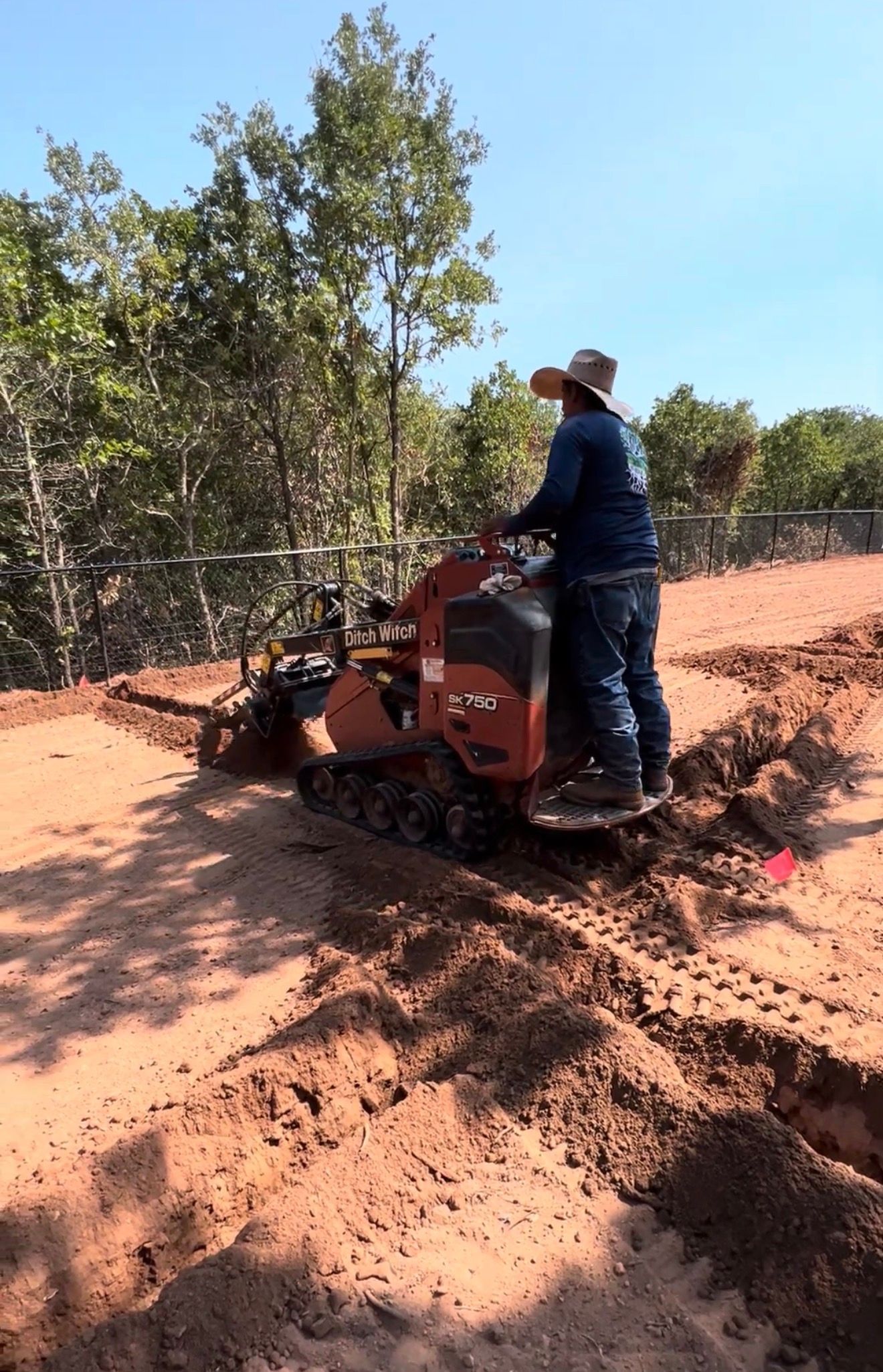 Person operating a red tiller on a sandy path beside trees under a clear blue sky