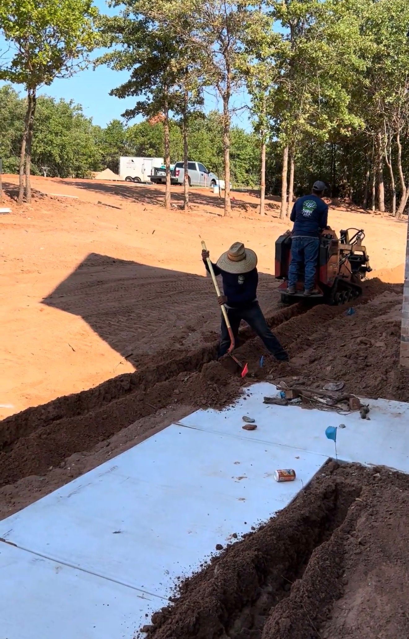Workers installing a white concrete walkway along a dirt road in a wooded area