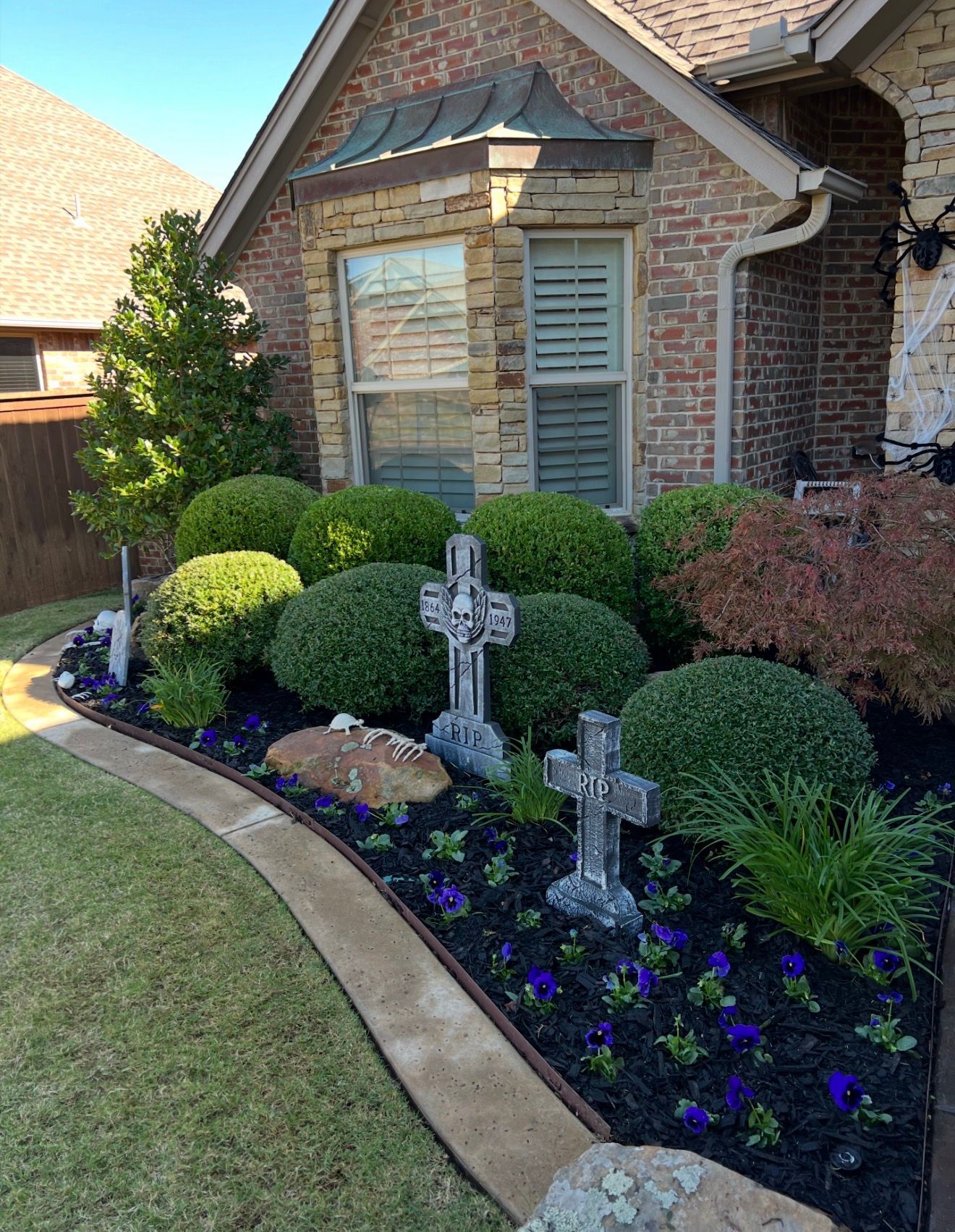 Brick house front garden with trimmed shrubs, purple flowers, and stone cross ornaments along a curved walkway
