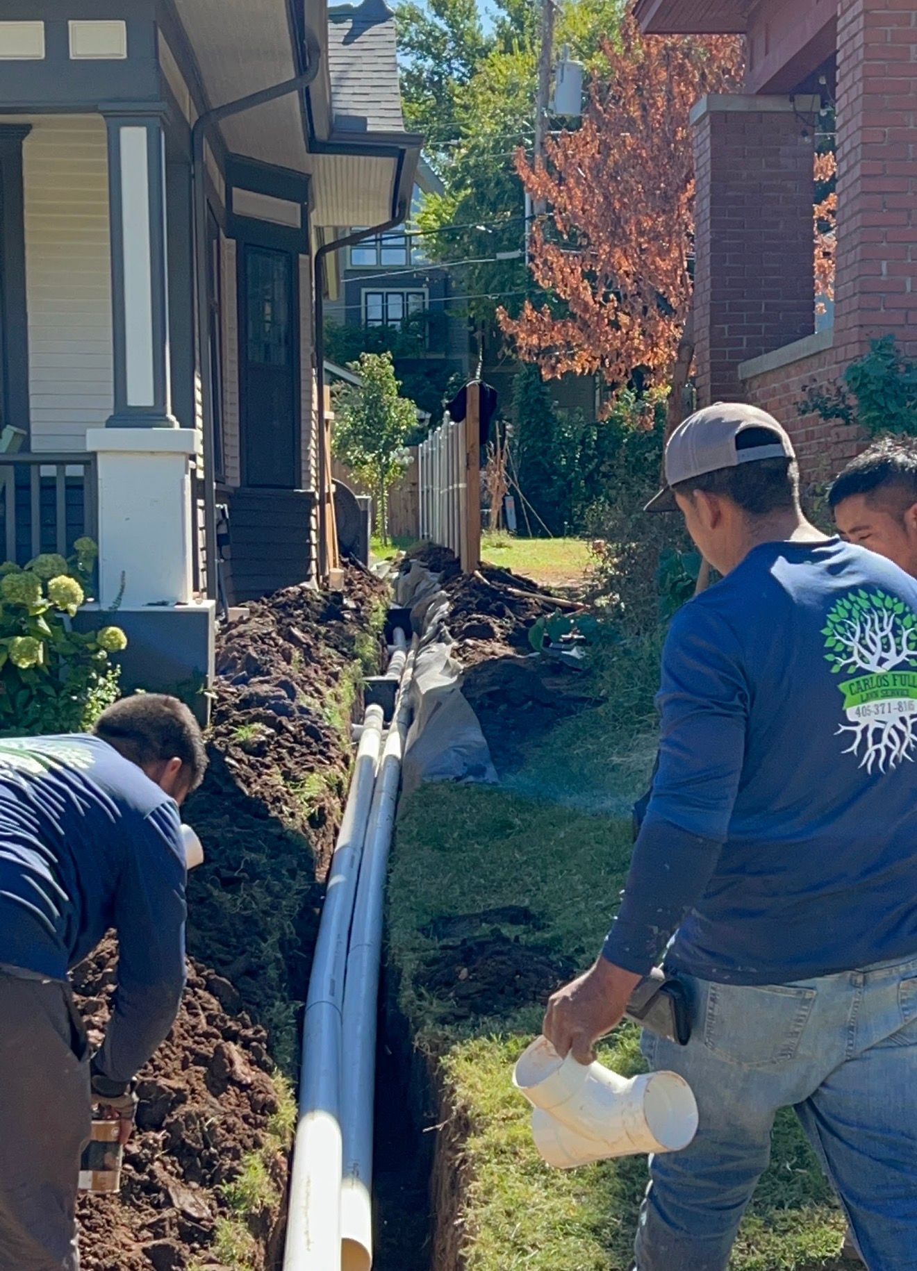 Workers installing drainage pipe along a narrow trench beside houses on a sunny day