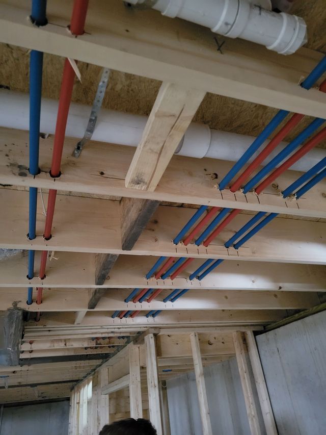 Exposed ceiling joists with white pipes and red and blue plumbing lines in a construction site