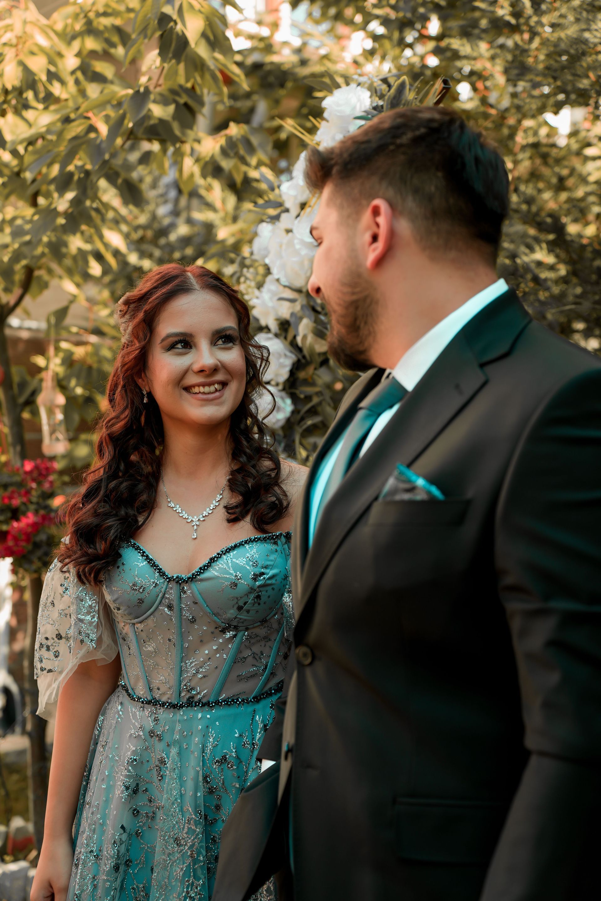 Couple dressed formally outdoors, smiling at each other under trees with white blossoms.