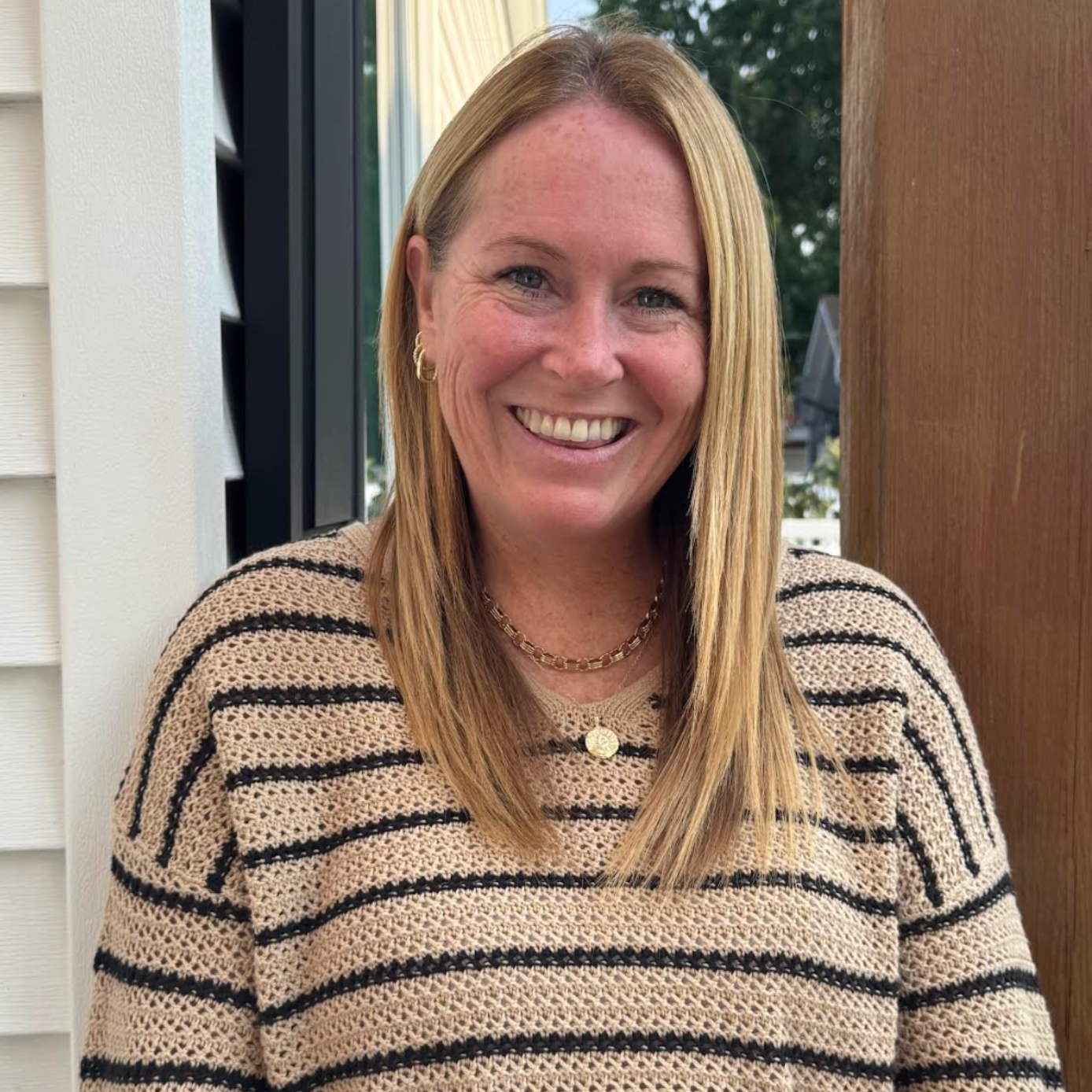 A woman in a grey sweater is smiling in front of a bush.