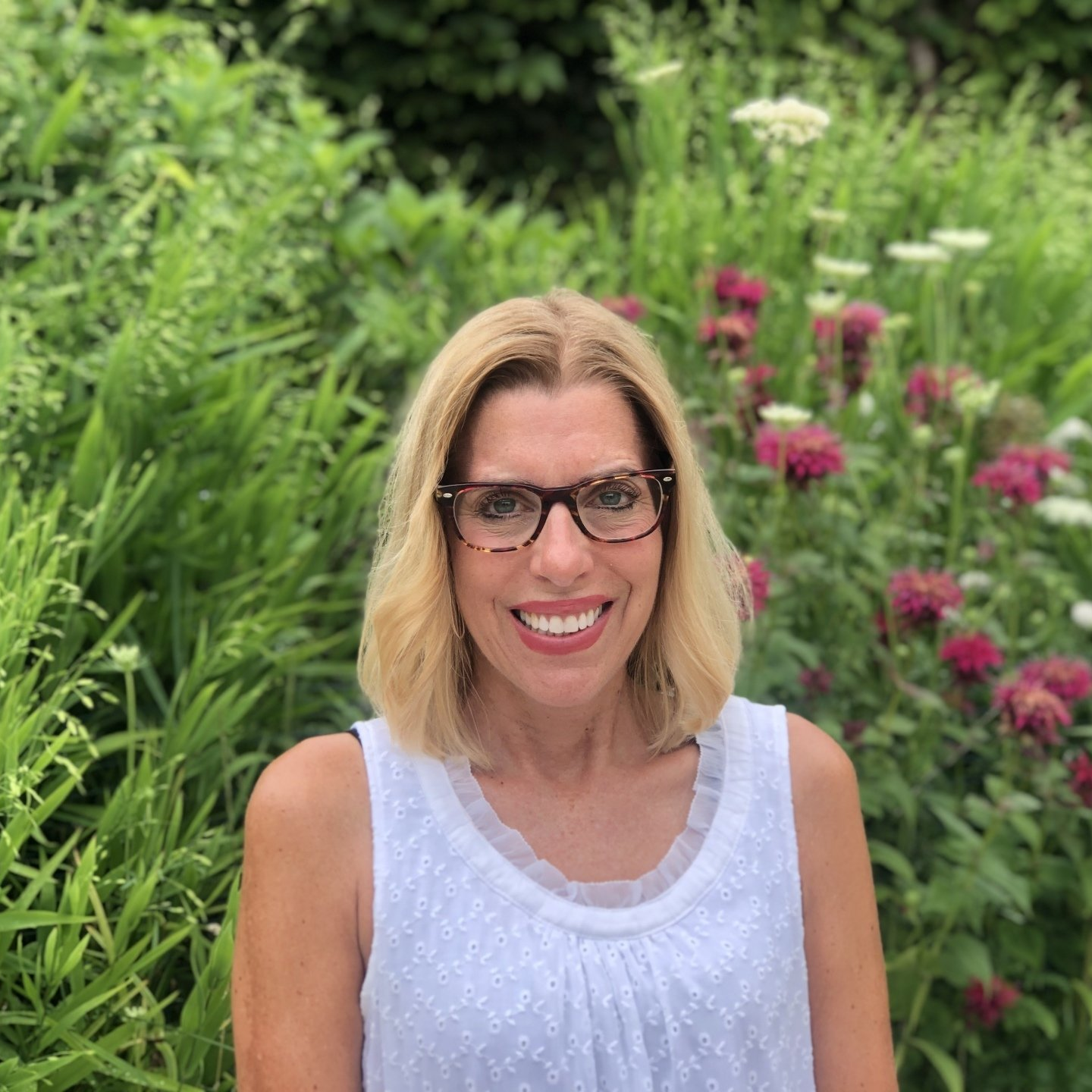 A woman wearing glasses and a white tank top is smiling in front of a bush.