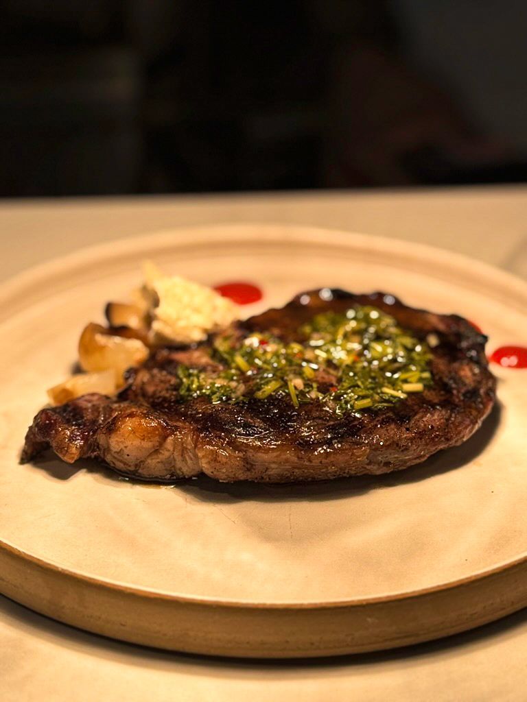 A grilled steak topped with chimichurri sauce, served with roasted garlic cloves on a minimalist ceramic plate.