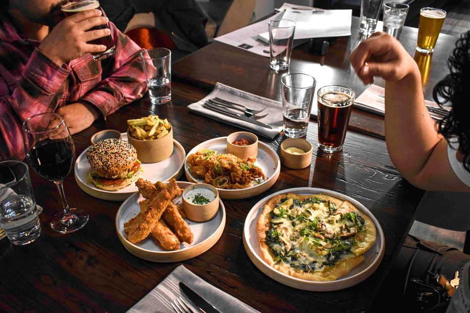 Dining table with several plates of food, drinks, and people’s hands around it in a restaurant.
