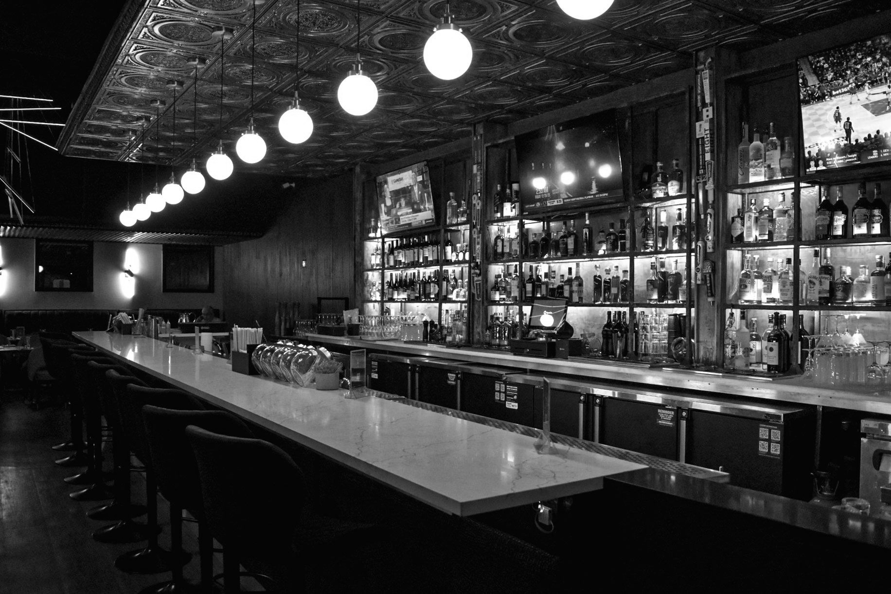Black and white photo of a bar interior with a long counter, stools, and shelves stocked with bottles.
