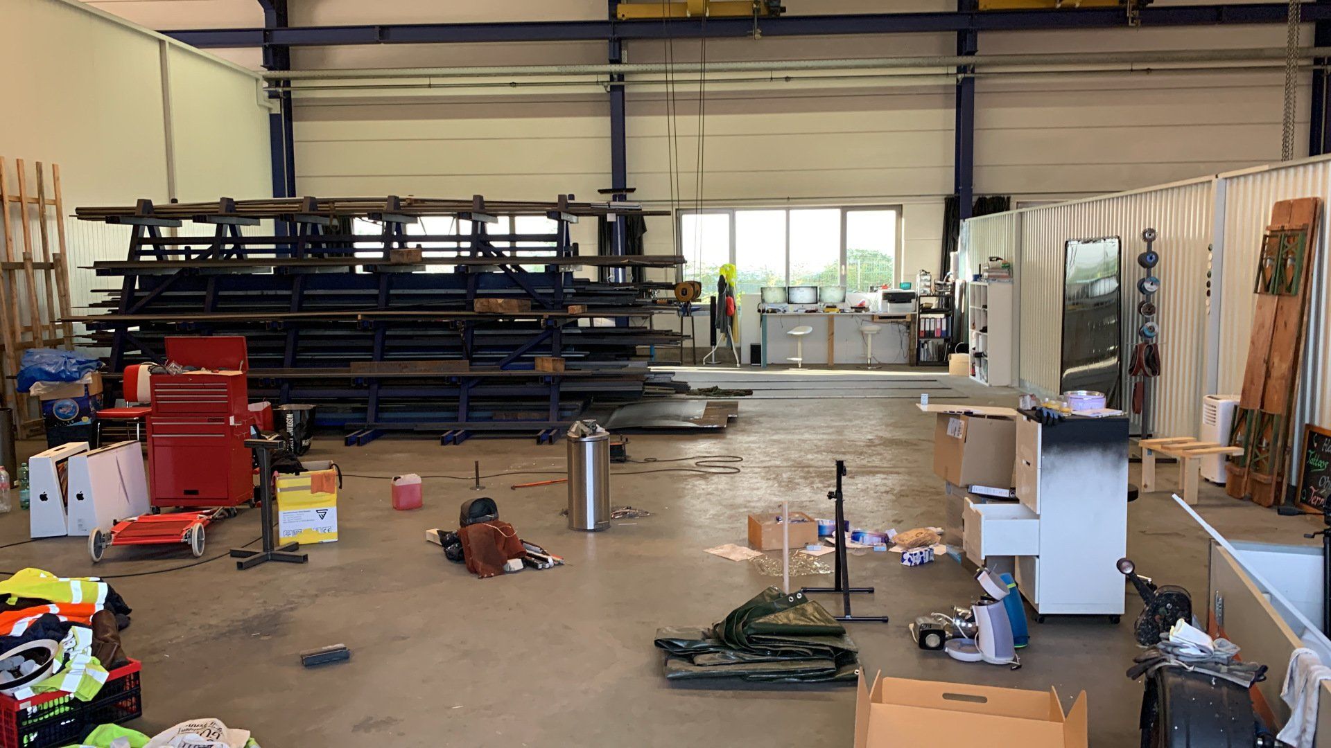 Interieur van een magazijn met metalen stellingen, gereedschap, dozen en een persoon bij een raam.