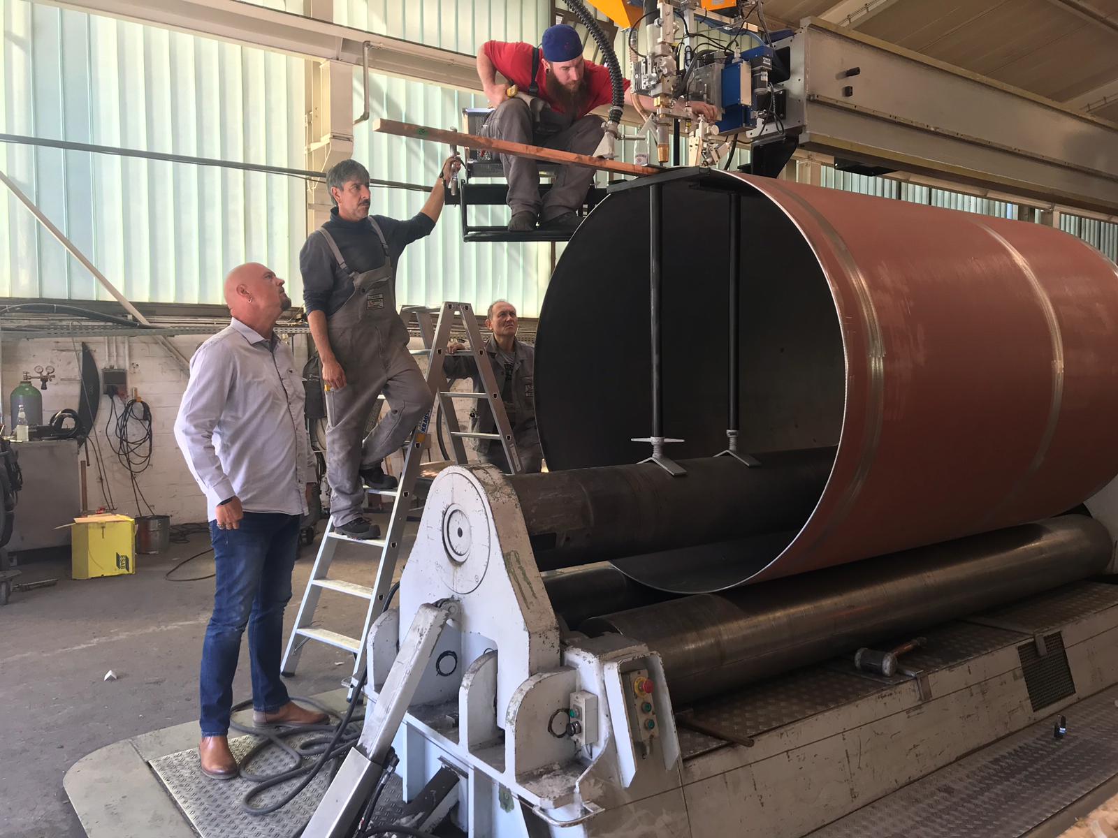 Arbeiders monteren een grote cilindrische tank in een werkplaats. Een man in een hemd kijkt toe.