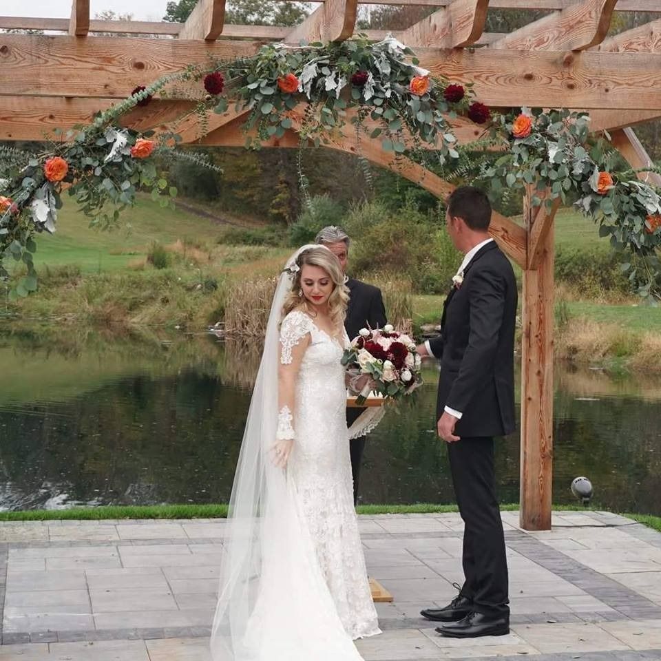 Bride and groom under a floral arch by a pond, standing on a stone patio outdoors